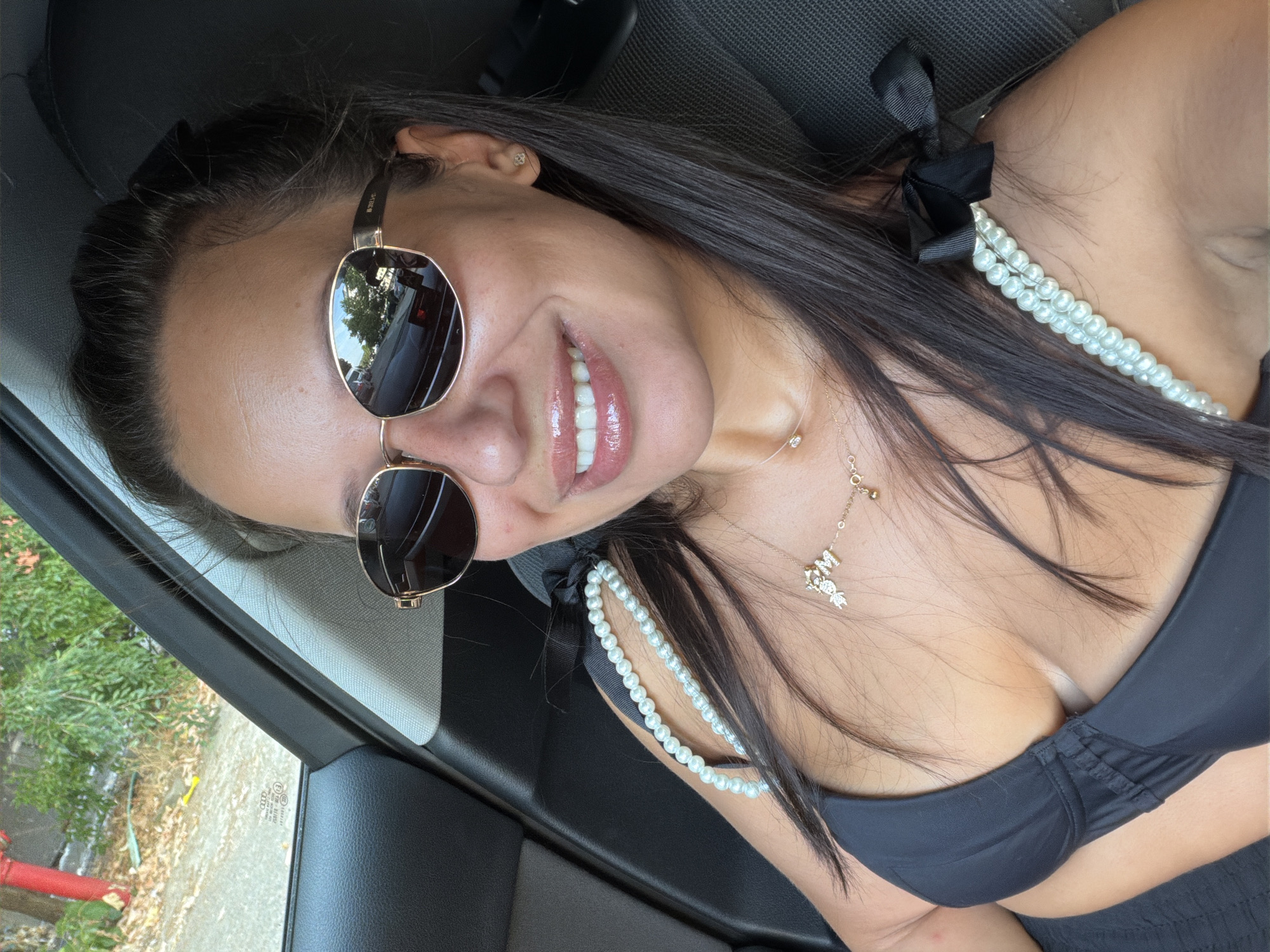 The image shows a woman with long dark hair, wearing a black bikini top, sunglasses, and a necklace. She is in the passenger seat of a car. The environment outside the car appears to be an urban street with trees and buildings. The photo was taken on July 25, 2023, at 12:36 PM in Burgas, Bulgaria.