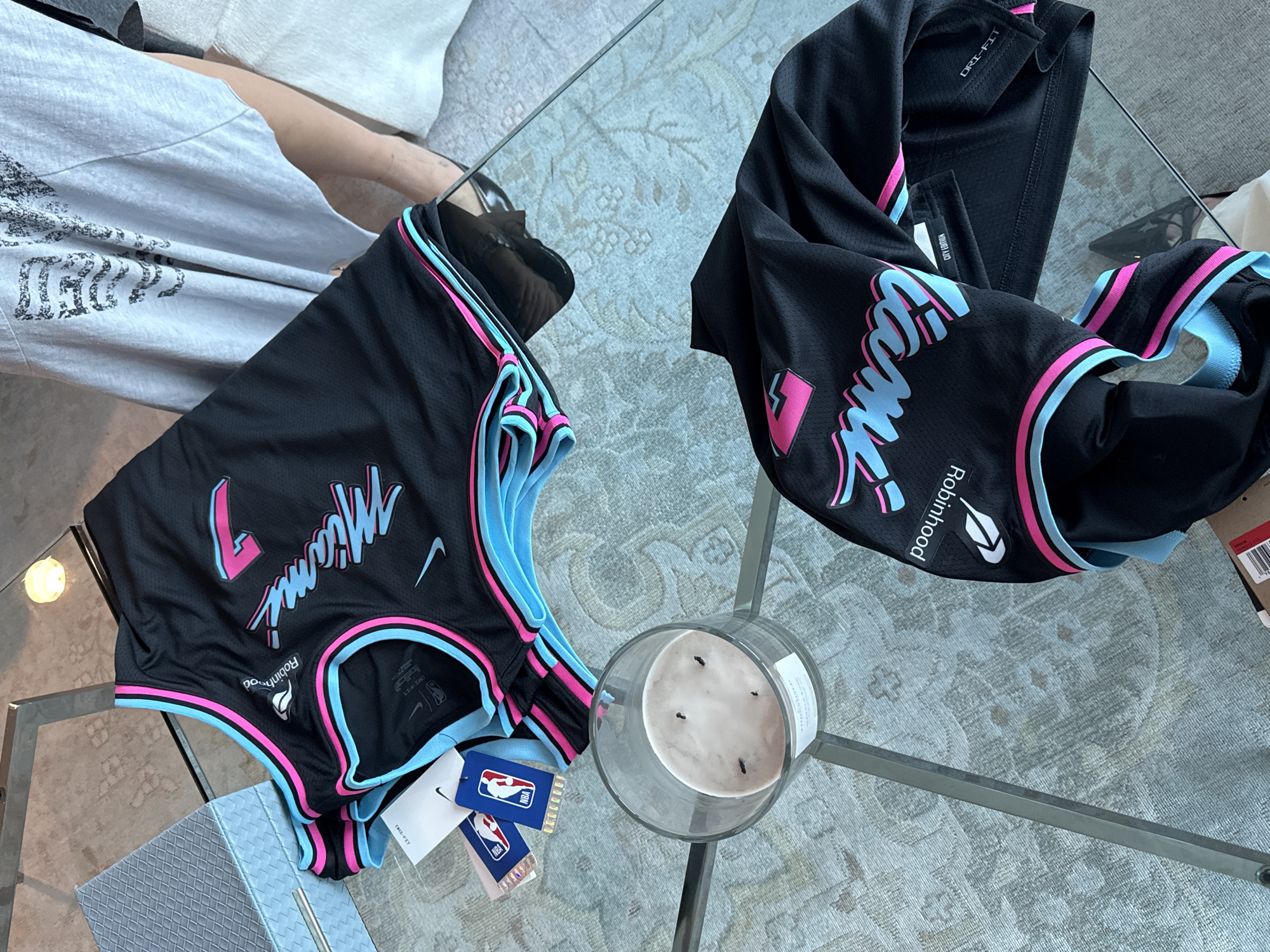 Two Miami Heat City Edition jerseys, likely new given the attached tags, are laid out on a glass coffee table. Each is black with "Miami" written across the chest in a teal and pink font, accompanied by the number "7" and a white Nike logo. A "Robinhood" sponsor patch is visible on both jerseys. The setting appears to be indoors, possibly a living room, with a person visible on the left in a grey shirt and the edge of a sofa in the background. A partially used candle sits between the jerseys on the table. There is no indication of the jerseys being located in the city of Miami, but their design alludes to the location.