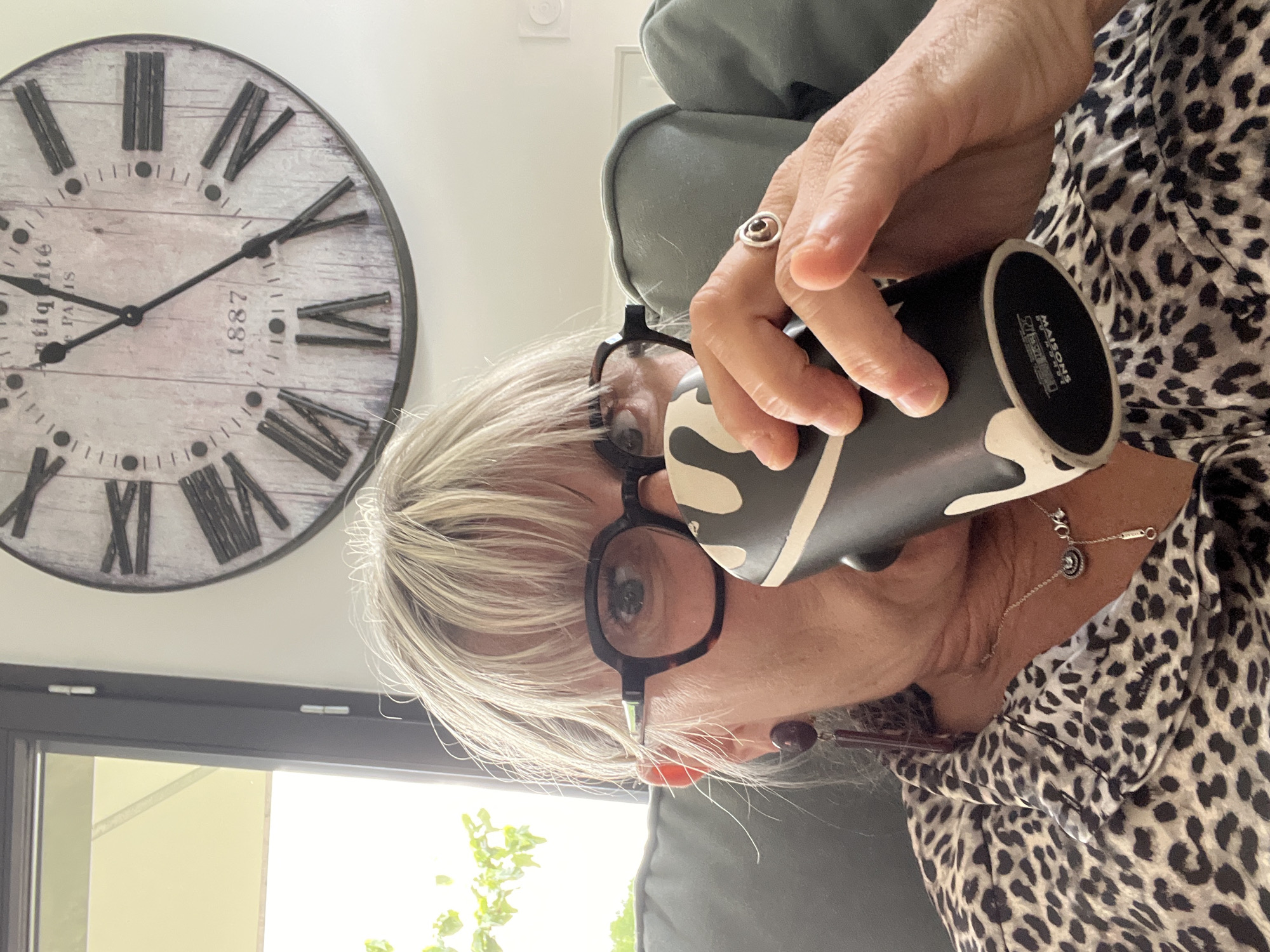 A middle-aged, fair-skinned woman with blonde hair and glasses is drinking from a black and white mug. She is wearing a leopard-print shirt and a ring on her finger. A clock with Roman numerals hangs on the wall behind her. The clock indicates the time is approximately 8:50. The image was taken in Chambœuf, France.
