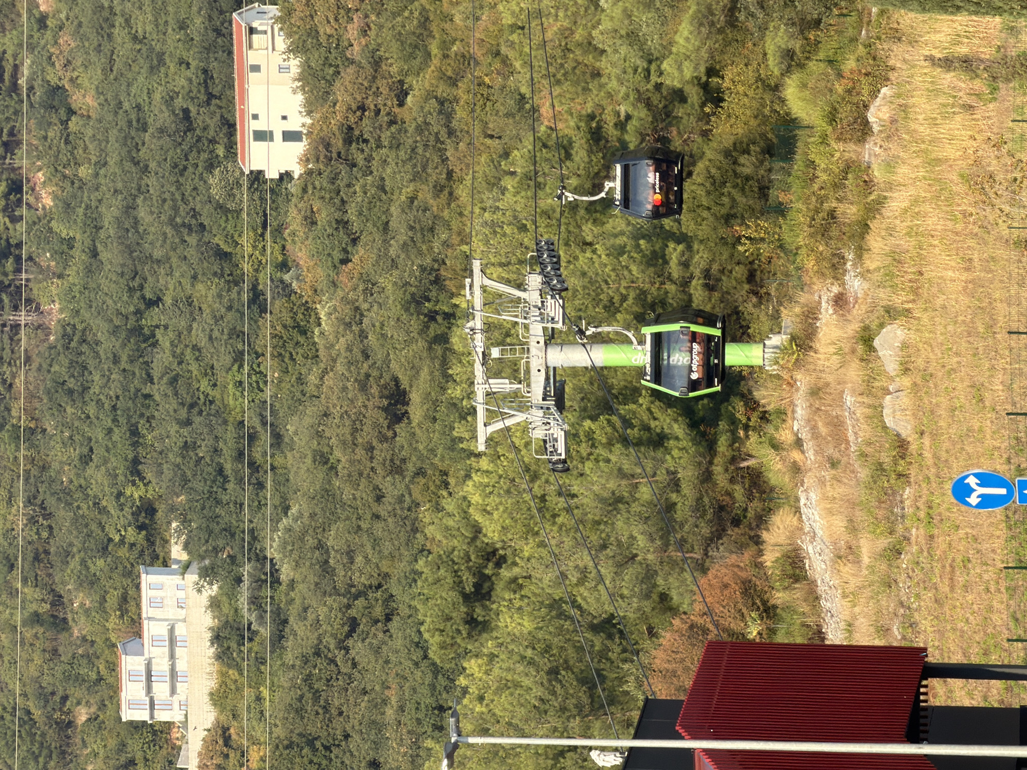 A daytime, medium-shot shows a cable car system with two cabins suspended above a hilly, tree-covered terrain in Dub, Montenegro. The cable cars are suspended from a grey metal support tower. The foreground features a modern red building with vertical ribbed siding on the left, and a street sign with arrows indicating a fork in the road. Dry, golden grass covers the slopes. In the background, two large, modern white houses are visible nestled among the trees on the hillside. One house is larger and on a higher elevation, featuring multiple stories and numerous windows. The other house is smaller and appears to be two stories with a red roof. The environment suggests a relatively warm climate with abundant vegetation.