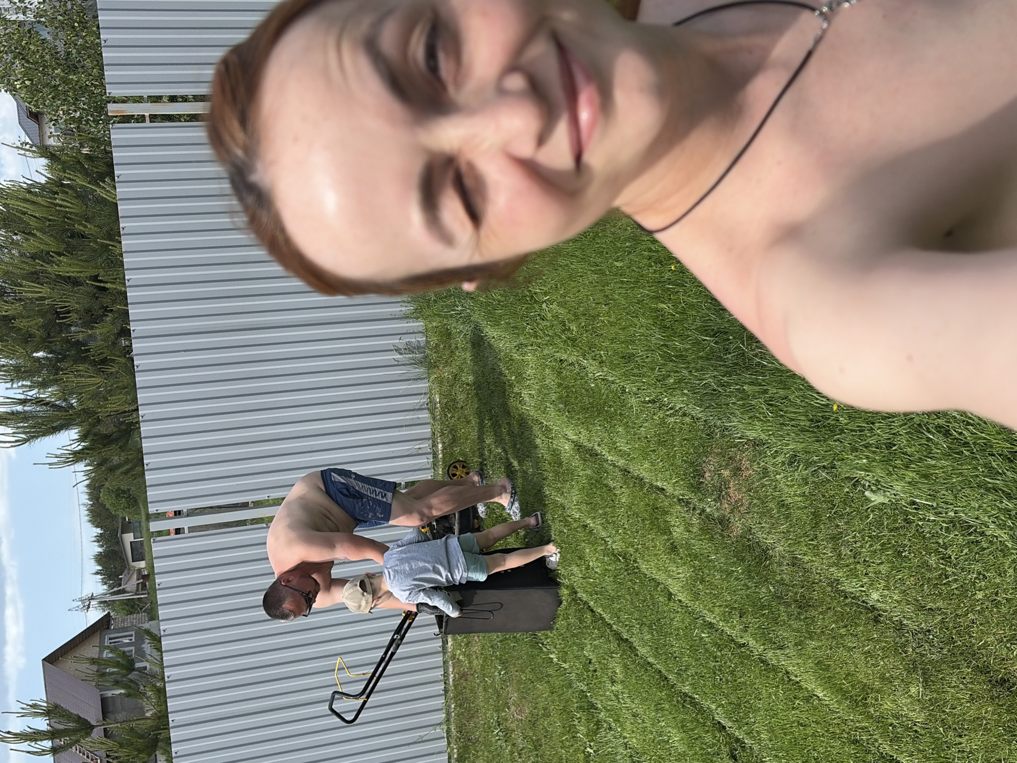 A woman holds a phone and is taking a selfie. Behind her, a man and child are pushing a lawnmower across a lawn near a gray metal fence. In the background is a building and trees. The location is Chaikovsky, Russia.