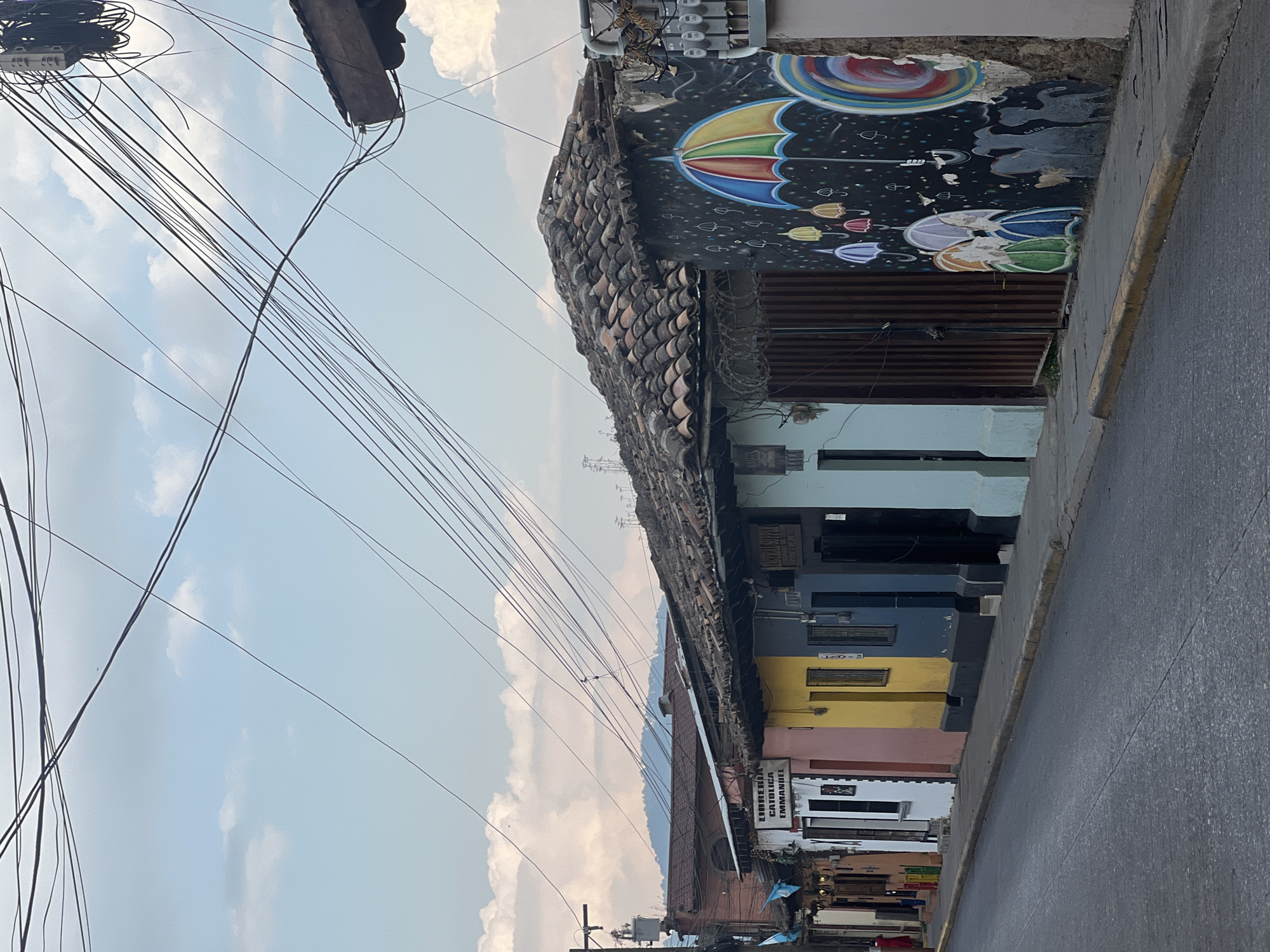 The image depicts a street scene in La Esperanza, Honduras. A row of colorful buildings lines the right side of the street, featuring hues of white, pink, yellow, blue, and light blue. The buildings have tiled roofs and a mural of umbrellas. Above, a dense network of electrical wires crisscrosses against a backdrop of a cloudy sky. A street runs along the side of the buildings.