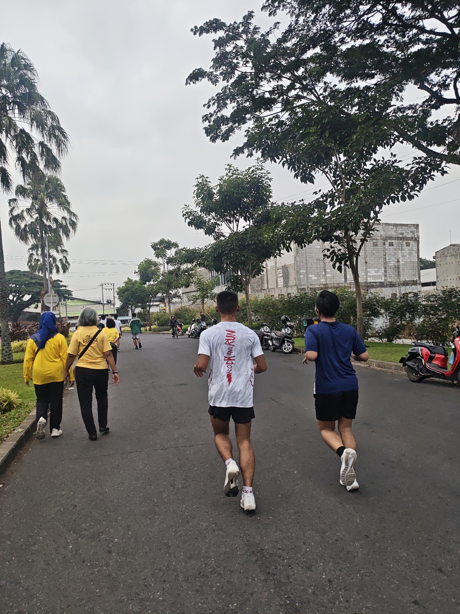 In the city of Bacin, Indonesia, on a paved road, a group of people are engaged in a running or walking activity. The day is overcast, with a pale, cloudy sky. Tall palm trees and other leafy trees line the right side of the road, casting shadows. In the background, several low-rise buildings with light-colored brickwork are visible, alongside power lines and poles. To the right of the road, several parked scooters are present, one of which is red and appears to be a modern scooter.
Three individuals are prominently featured in the foreground, seen from behind. The person closest to the camera on the right is wearing a white t-shirt with red lettering that reads "ROCK and RUN," black shorts, and white running shoes. To their left, another person is wearing a dark blue t-shirt, black shorts, and white running shoes with black socks. Further to the left, two women are walking ahead. The woman in the middle is wearing a yellow t-shirt with a logo that says "BUDYA" and black pants, and the woman to her left is wearing a bright yellow long-sleeved shirt and black pants, with a blue headscarf. Several other people are visible further down the road, including a person in a green shirt running. The activity appears to be a group fitness event or a casual recreational outing.
