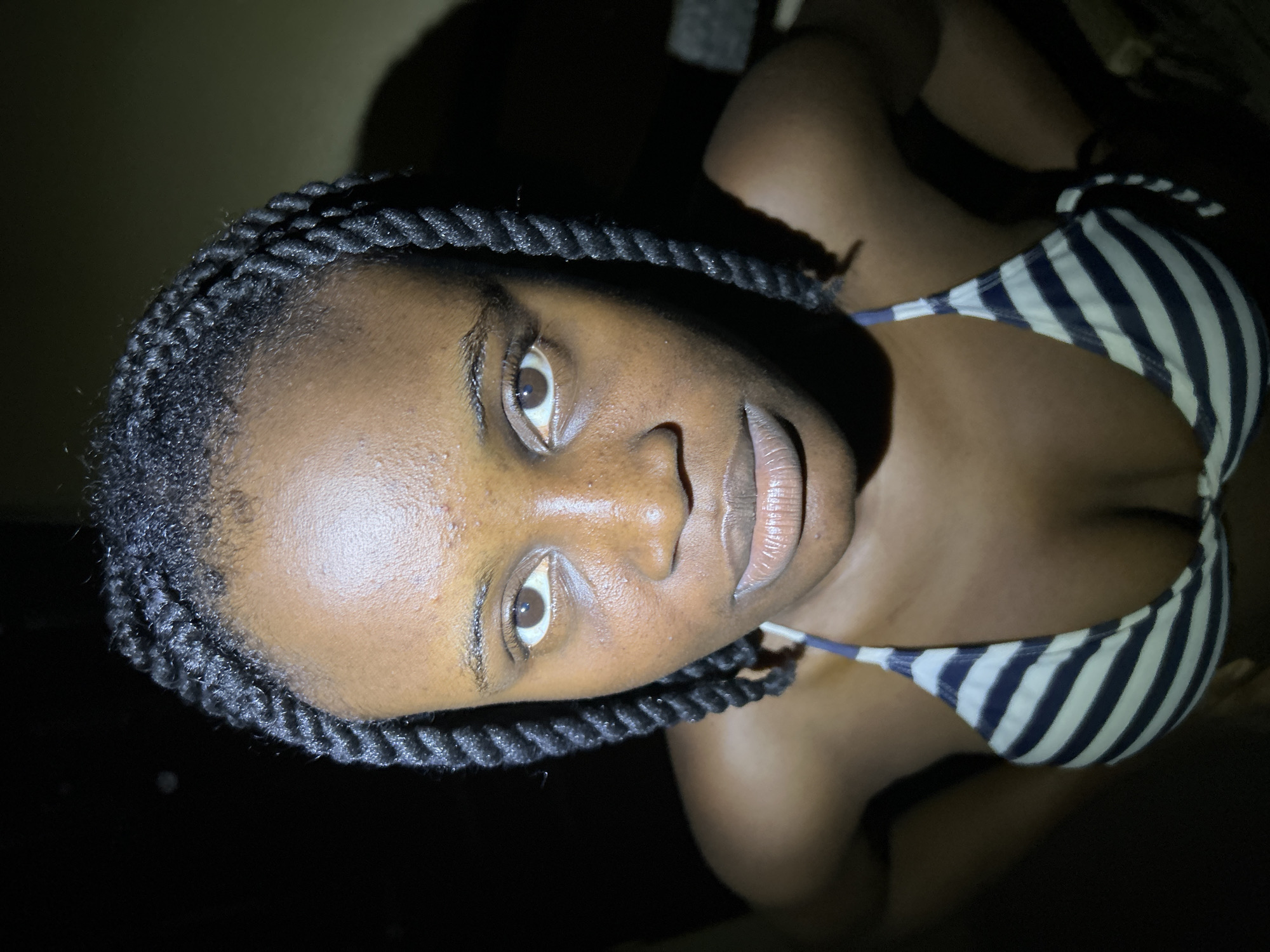 The image is a close-up, high-angle shot of a young Black woman, seemingly in an outdoor setting at night. She has dark skin, braided hair, and is wearing a striped bikini top. Her face is the primary focus, with her eyes looking directly at the camera. The lighting appears to be artificial and from above, casting shadows on her face and highlighting her skin. The background is dark and out of focus, with some hints of a wall or structure. There is no indication of the specific location within Ihiagwa, Nigeria.
