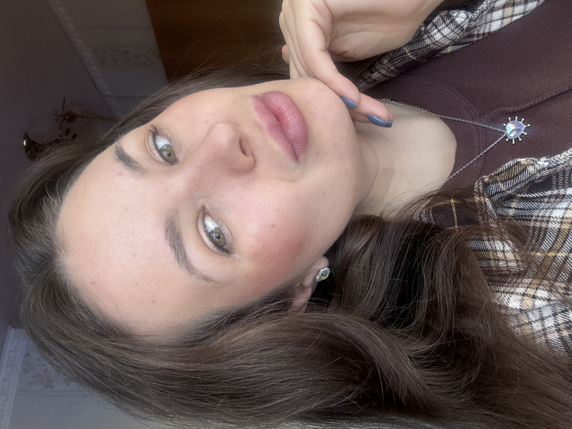 A young woman with brown hair and light green eyes is looking upwards toward the camera. She has on light pink lipstick and blush. She is wearing a maroon colored shirt with a plaid jacket on top. She has a silver necklace with a heart shaped pendant and silver earrings. The setting and location is unknown, could not confirm city of Bely Yar, Russia.