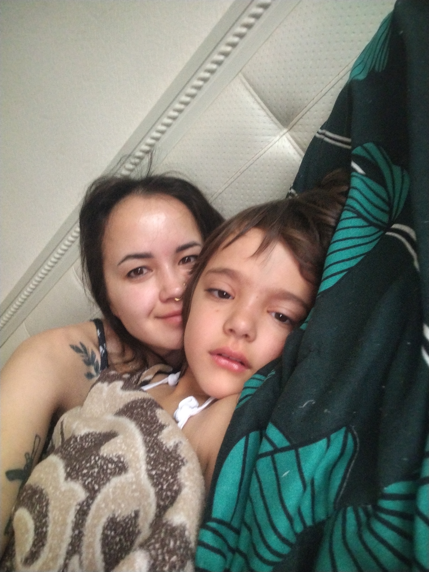 Two individuals, likely a young woman and a child, are indoors, possibly in a bed. The woman is positioned behind the child, her face visible and turned slightly towards the camera. The child appears partially covered by a patterned blanket and a dark-colored fabric with green designs. The background includes a padded headboard. No details indicating the location as Noviy, Russia are visible.