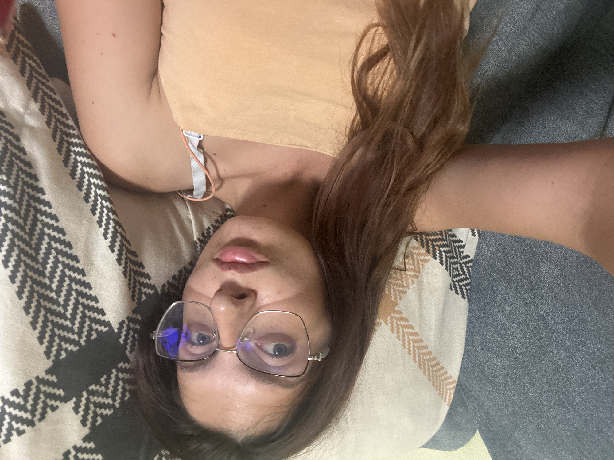 A young woman with long brown hair and blue eyes is lying on a sofa. She is wearing glasses and a peach-colored tank top. The sofa is covered with a blue textured fabric on one side and a patterned throw blanket on the other. The throw blanket has a geometric design in beige, black, and white. The woman is looking towards the camera with her lips slightly parted. The scene appears to be indoors, possibly a living room. The specific location in the city of Tryavna, Bulgaria is not determinable from the image alone.