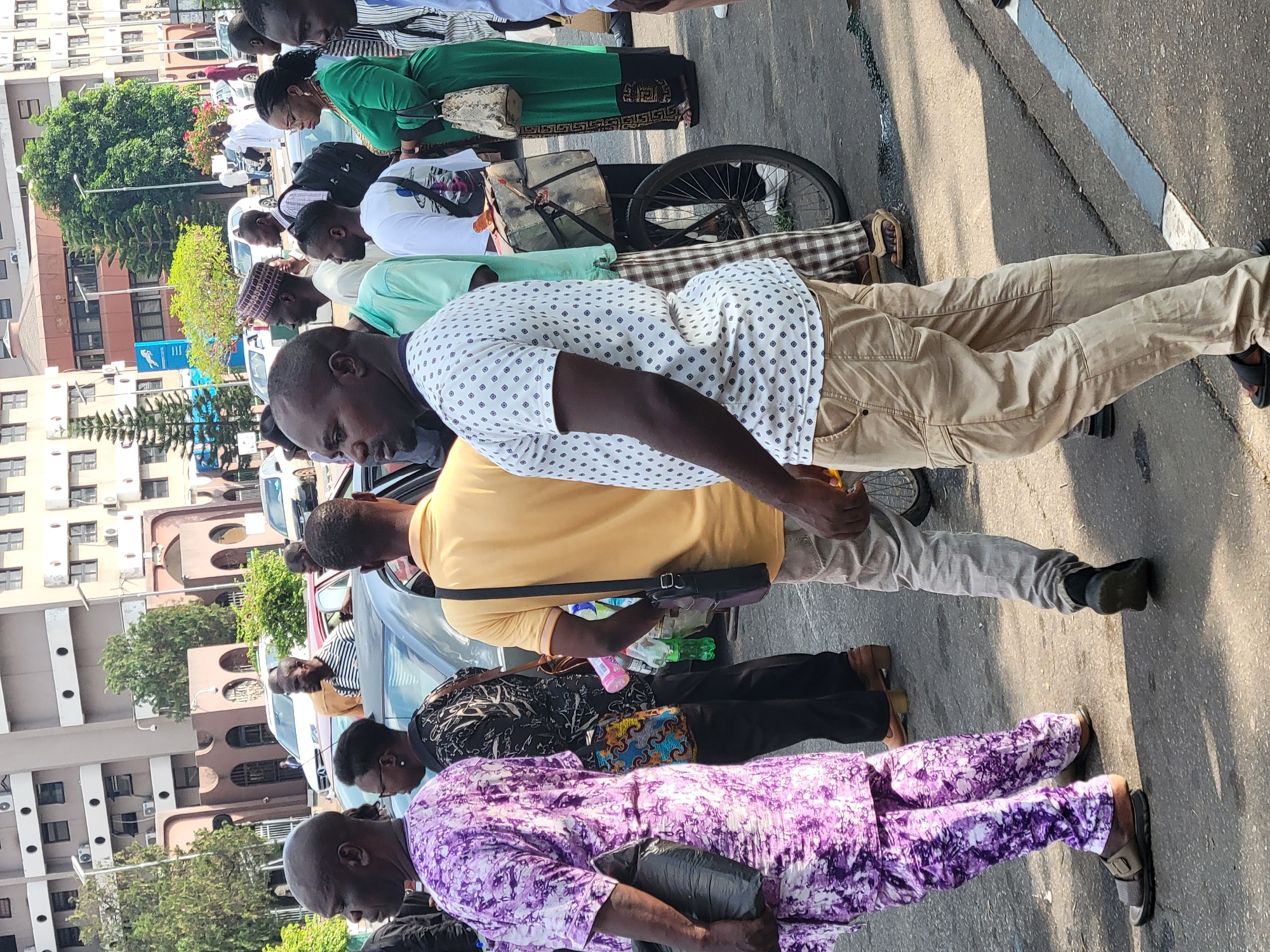 The image shows a group of people standing on a street in Abuja, Nigeria. The people are dressed in a variety of casual and traditional clothing. Some are standing, while others are hunched over. There are several cars parked along the street. There are buildings and trees visible in the background. A bicycle is visible in the image.