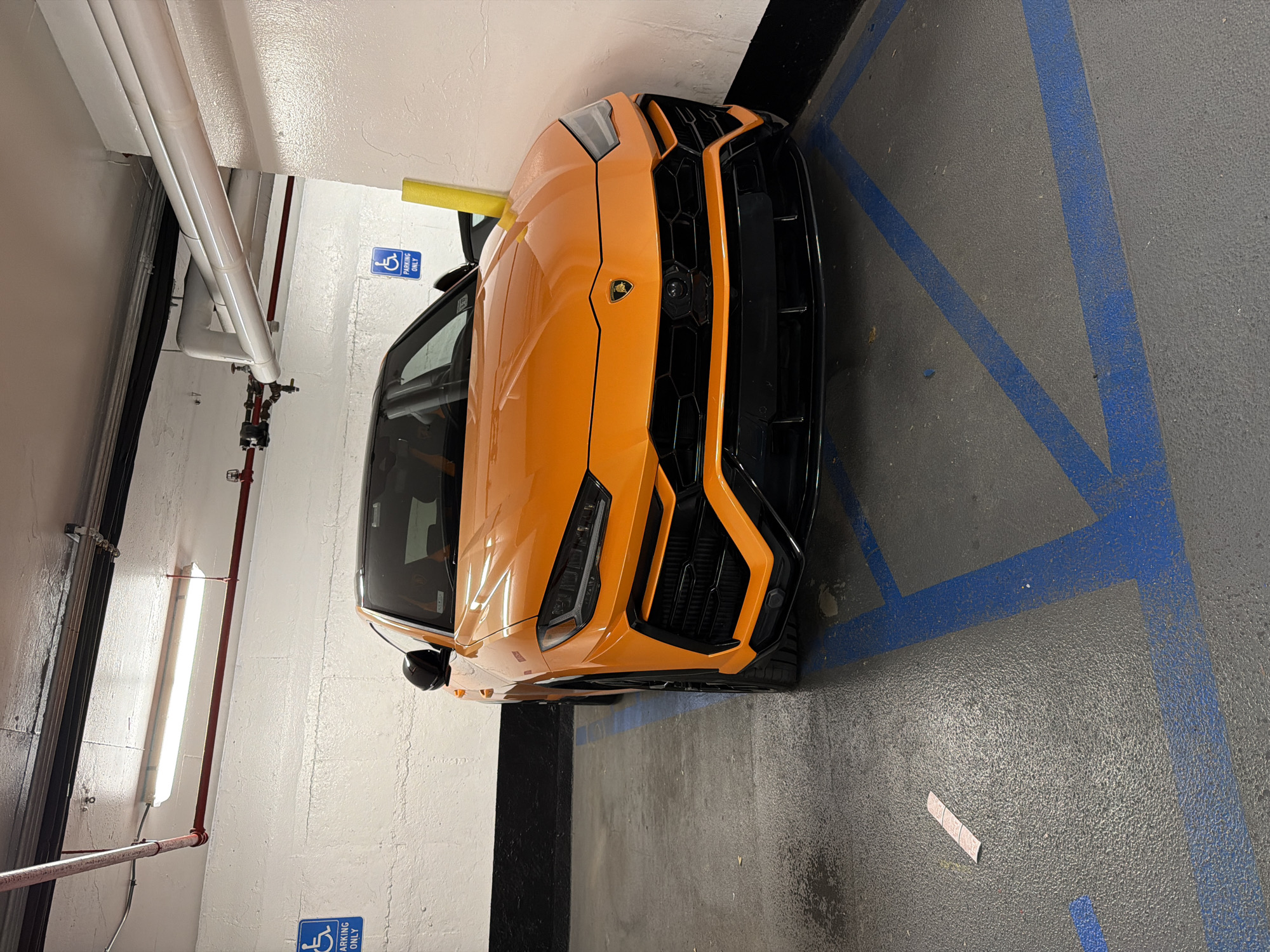 An orange Lamborghini Urus is parked in a handicapped parking spot in an underground parking garage. The car is facing the viewer, and its front end is the main focus of the image. The parking garage has a concrete floor with blue painted lines marking the parking spaces, and white painted walls. There are also pipes and lighting fixtures visible on the ceiling of the garage. The handicapped parking sign is visible on the wall behind the car. A yellow safety pole is positioned next to the car.