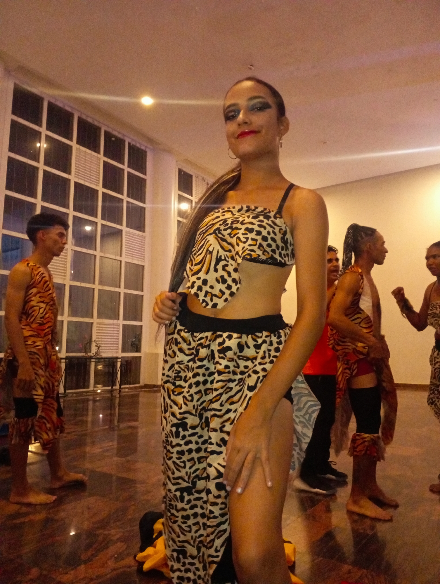 A group of people are inside a building with a marble floor. The focus is on a woman in the center, wearing a leopard-print two-piece outfit with black trim. Her hair is pulled back in a ponytail and she wears heavy eye makeup and red lipstick. She is looking towards the viewer and smiling. In the background, other individuals stand in what appears to be similar tribal-themed outfits, some resembling tiger stripes. A window with square panes is also visible in the background. The lighting in the room is dim. The location is Pedro González, Venezuela.