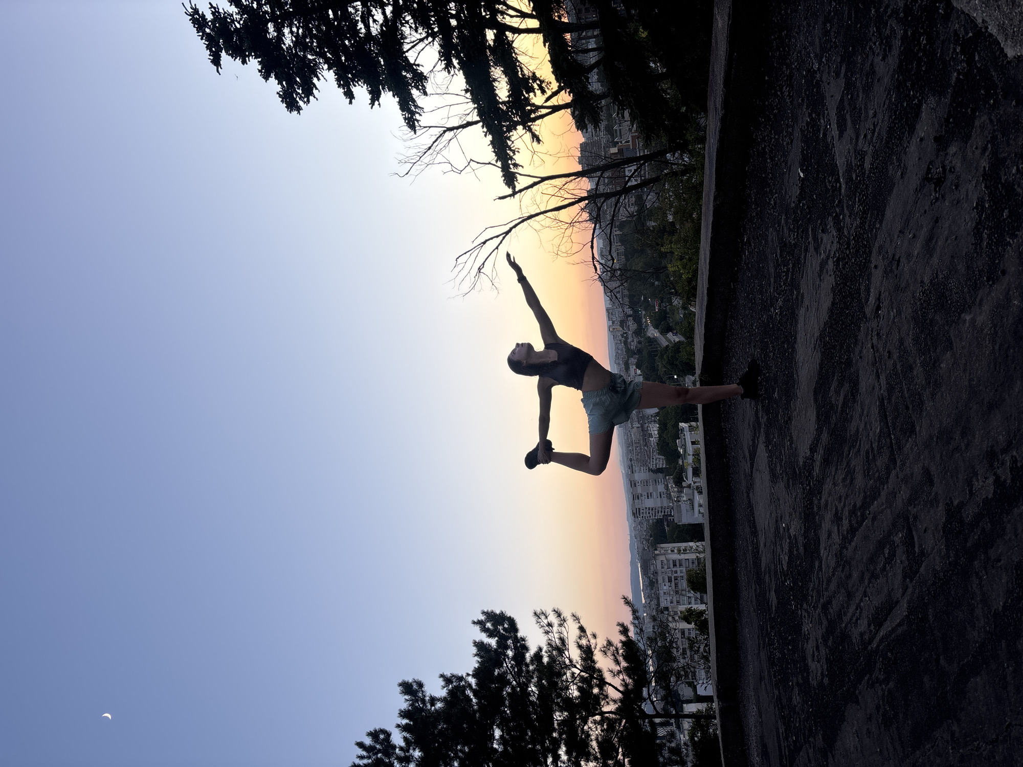 A young woman is performing a yoga pose, "Dancer's Pose" (Natarajasana), on a rooftop overlooking the city of Varna, Bulgaria, at dusk. She is silhouetted against a gradient sky transitioning from a clear blue at the top to soft oranges and pinks near the horizon. The city skyline, with its multi-story buildings, is visible in the background. She is balancing on her left leg with her right leg bent and held by her right hand, while her left arm is extended forward. She wears a dark crop top and light-colored shorts. To the left and right of the frame, the dark silhouettes of pine trees frame the scene. A sliver of a crescent moon is visible in the upper left corner of the sky. The rooftop surface is dark and textured, appearing to be asphalt or a similar material. The overall mood is serene and athletic, capturing a moment of calm amidst the urban landscape as day transitions to night.
