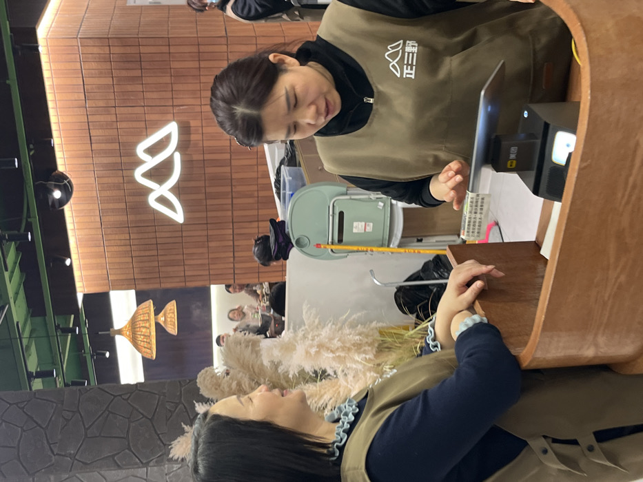 This image depicts an interior scene within a Zheng San Ye restaurant, located in Jiangjin, China. In the foreground, two adult females, identified as employees by their identical khaki-colored aprons bearing the "正三野" mountain logo, stand behind a wooden service counter. The woman on the left, with dark hair, wears a dark blue long-sleeved shirt with light blue ruffled cuffs and smiles as she looks towards the right. Her hands rest on the counter's edge. The woman on the right, also with dark hair, wears a black long-sleeved shirt and is focused downwards, her right hand positioned over a tablet device mounted on a black payment terminal displaying the Meituan logo.
The background shows a dining area with several indistinct figures seated at tables, consistent with patrons. Tall, light-colored pampas grass plumes are visible as decoration. The left wall features a dark, textured stone-like finish, while the right wall is covered in reddish-brown rectangular tiles with a prominent white neon outline of the mountain logo. The ceiling is green, equipped with track lighting and two woven, dome-shaped pendant lamps. Behind the counter area, a light-green utility bin, a yellow pole, and black trash bags are discernible.