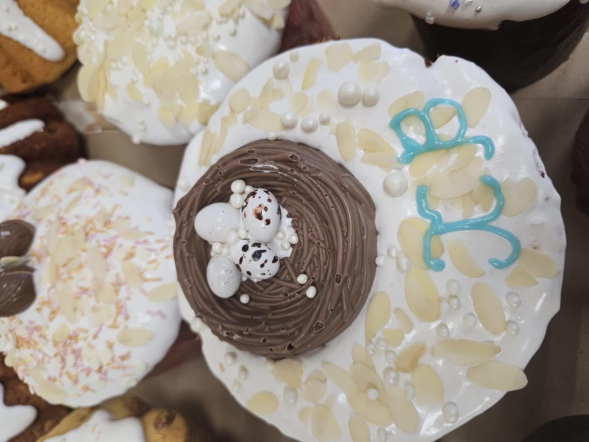 The image shows a high-angle, close-up view of several decorated Easter cakes, also known as kulich, arranged together. The cakes are topped with white icing and various decorative elements.
The central kulich features a chocolate nest in the center with three small decorative eggs nestled inside. The white icing around the nest is adorned with sliced almonds, small white pearl-like candies, and a teal-colored piped design of the letters "XB," which are Cyrillic initials for "Christ is Risen," a common Easter greeting.
Adjacent kuliches in the scene are decorated with similar white icing, almond slices, colorful sprinkles, and chocolate pieces.
Given the traditional nature of the cakes and the Cyrillic lettering, it is likely these cakes were photographed in Russia. As a common symbol associated with Orthodox Easter, it is plausible that the image could have been taken in Moscow, Russia, given its cultural significance there.