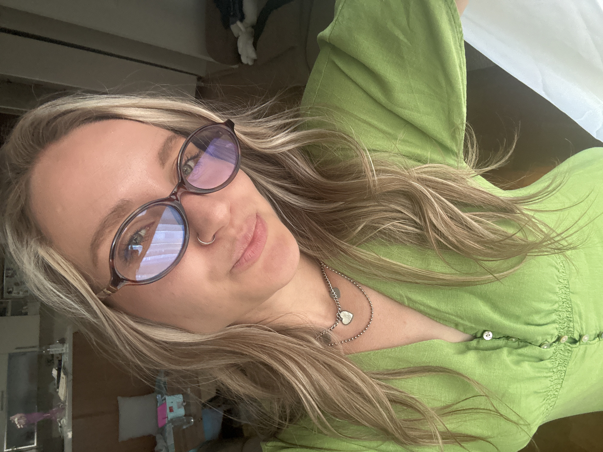 A young woman with blonde hair and glasses is posing for a selfie in a room. She is wearing a green dress and a necklace. The background shows a cluttered room with furniture and personal belongings. The image appears to have been taken indoors, possibly in a residential setting. There is no indication of the city of Esenyurt, Turkey, or any specific location within it.