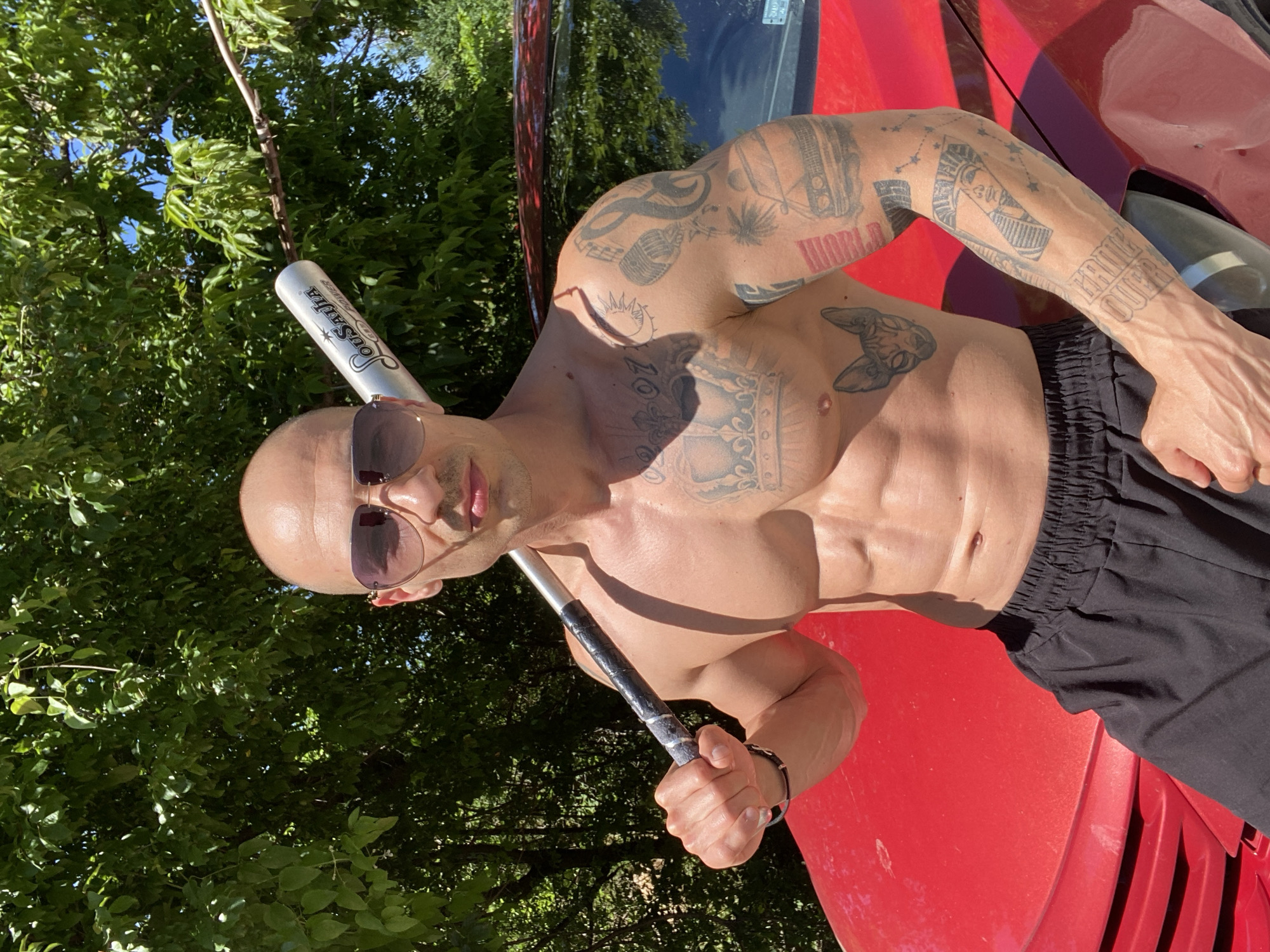 A shirtless, bald man wearing sunglasses is leaning against the hood of a red car outdoors. He holds a silver and black baseball bat. The man has numerous tattoos on his chest and arms. The background includes green trees and foliage. The location is not discernible as Pleven, Bulgaria, from the image alone.