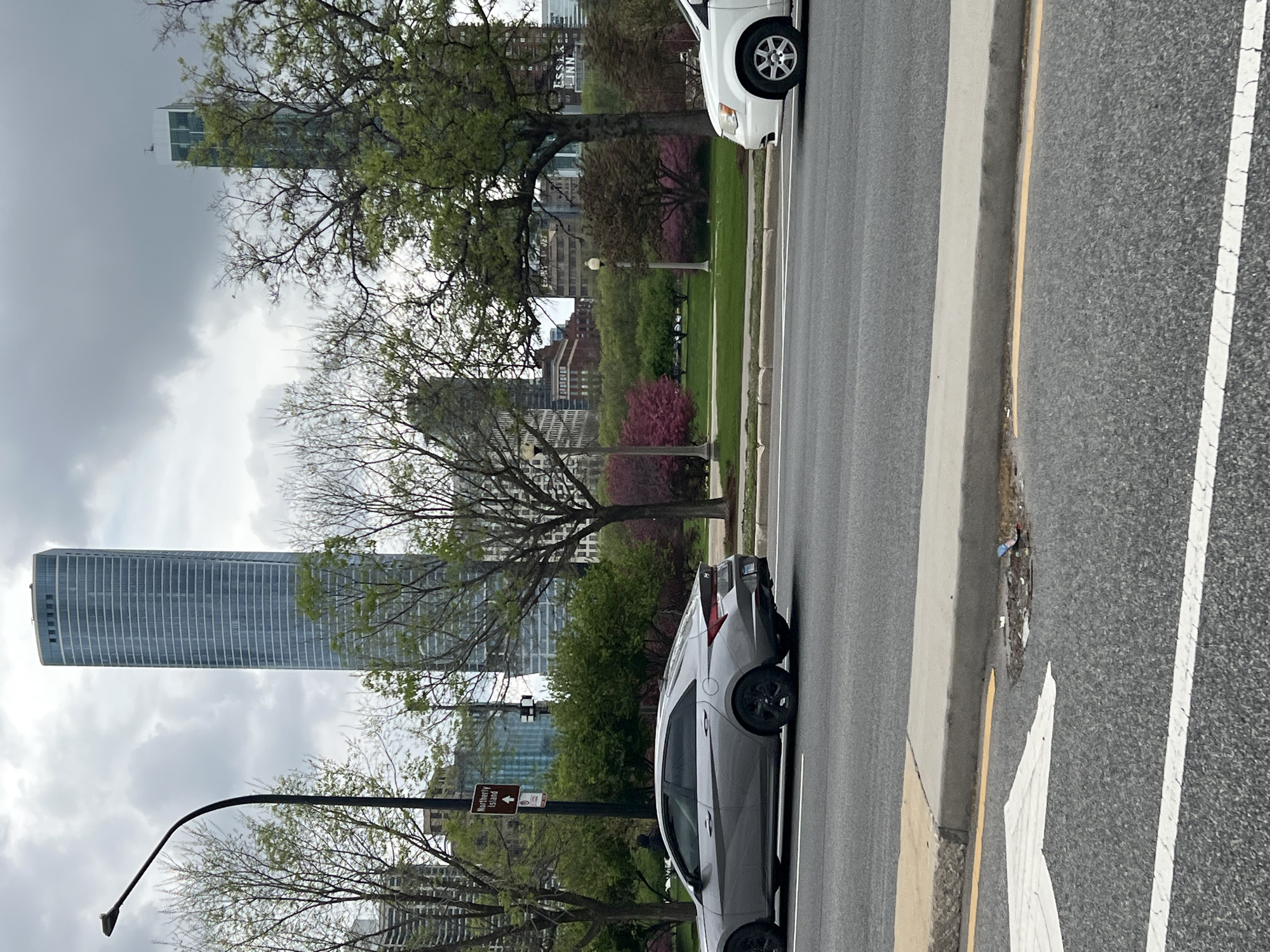 The image shows a street scene in Chicago, Illinois, USA. A silver car is parked alongside the curb on the left side of the street. A white car is partially visible on the right side of the street. In the background, there is a park with trees and flowering bushes. Buildings and skyscrapers are visible behind the park. The sky is overcast. A street lamp and a street sign indicating "Northerly Island" are visible near the silver car. The road is asphalt with painted lane markings.