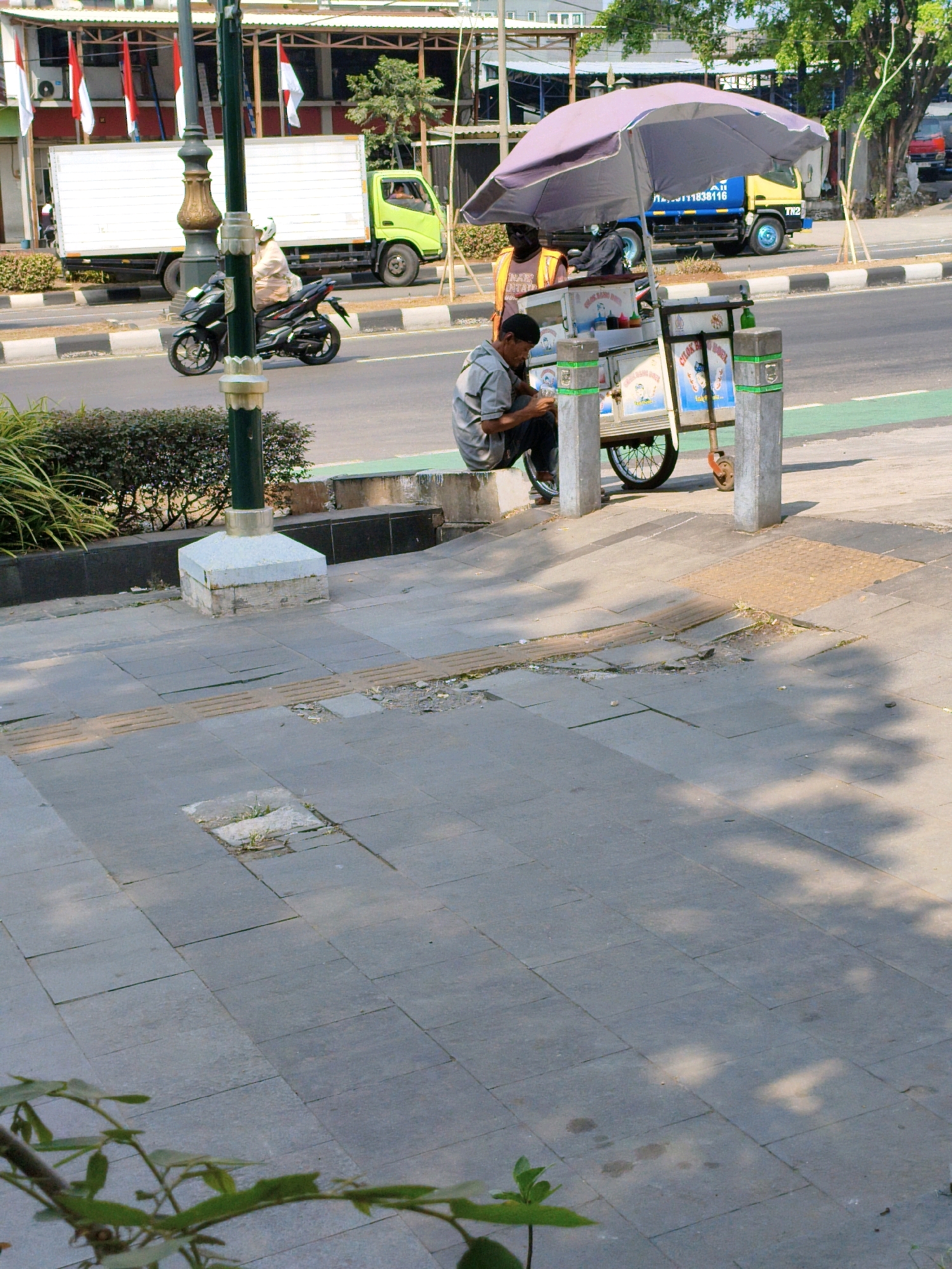 In Sukaraja, Indonesia, a street scene unfolds with a focus on a sidewalk area adjacent to a road. A person is seated on a concrete curb, engrossed in their mobile phone. Nearby, a food cart, equipped with a large purple umbrella, is positioned, suggesting a vendor's stall. The cart is laden with items, and graphics are visible on its side, indicative of its purpose. A yellow and blue truck is parked behind the food cart, partially obscured. The surrounding environment includes a lamppost with an ornate base, a concrete walkway with textured tactile paving for the visually impaired, and various plants and bushes. In the background, a white truck is seen on the road, with a rider on a black motorcycle passing it. The overall scene depicts a slice of everyday life on a street in an Indonesian city.