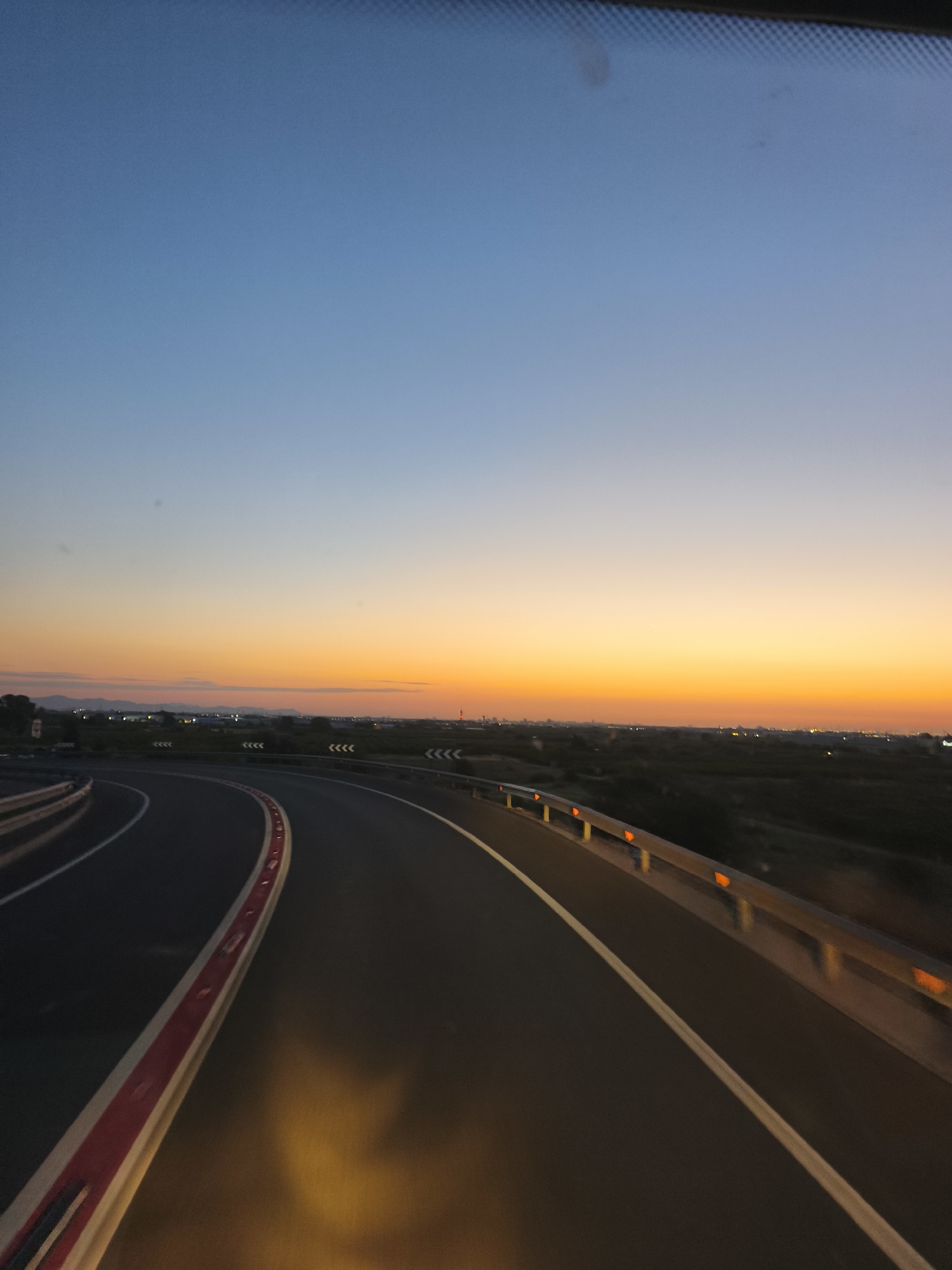 The image depicts a highway with a clear horizon at dusk. The sky transitions from a clear blue at the top to a vibrant orange and yellow near the horizon. In the distance, a city skyline with lights is visible. The highway itself is asphalt with white and red markings. Guardrails line the side of the road, and some have orange reflectors. The scene appears to be a view from a vehicle traveling on the highway. There are no people or animals visible in the image. The location is identified as the city of Alcàsser, Spain, likely on a highway leading into or out of the city. The time of day is clearly sunset or twilight.