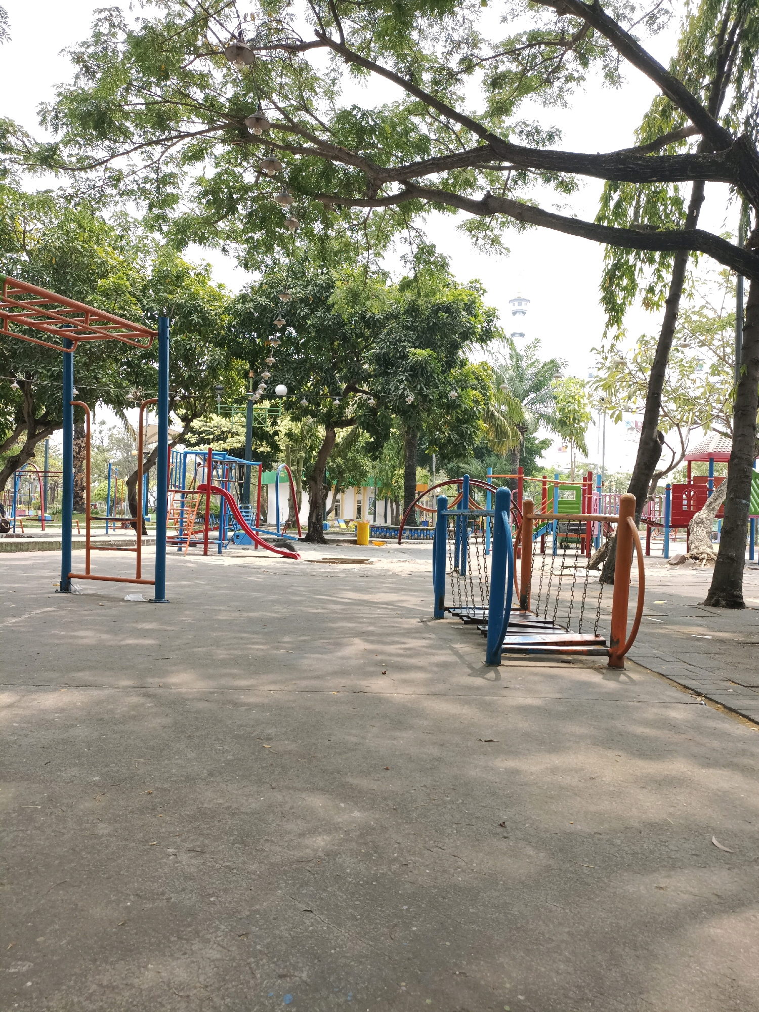 The image depicts a deserted playground in Lamongan, Indonesia, on a sunny day with a clear sky. The playground features various colorful structures, including monkey bars, slides, swings, and climbing frames, all constructed from metal and plastic materials. The ground is paved with concrete, and scattered leaves and debris are visible. Tall trees with lush green foliage surround the playground, casting dappled shadows on the ground. In the background, a building and what appears to be a Ferris wheel are visible, suggesting the presence of a larger recreational area or amusement park. The scene is peaceful and quiet, with no people present, creating a sense of solitude.