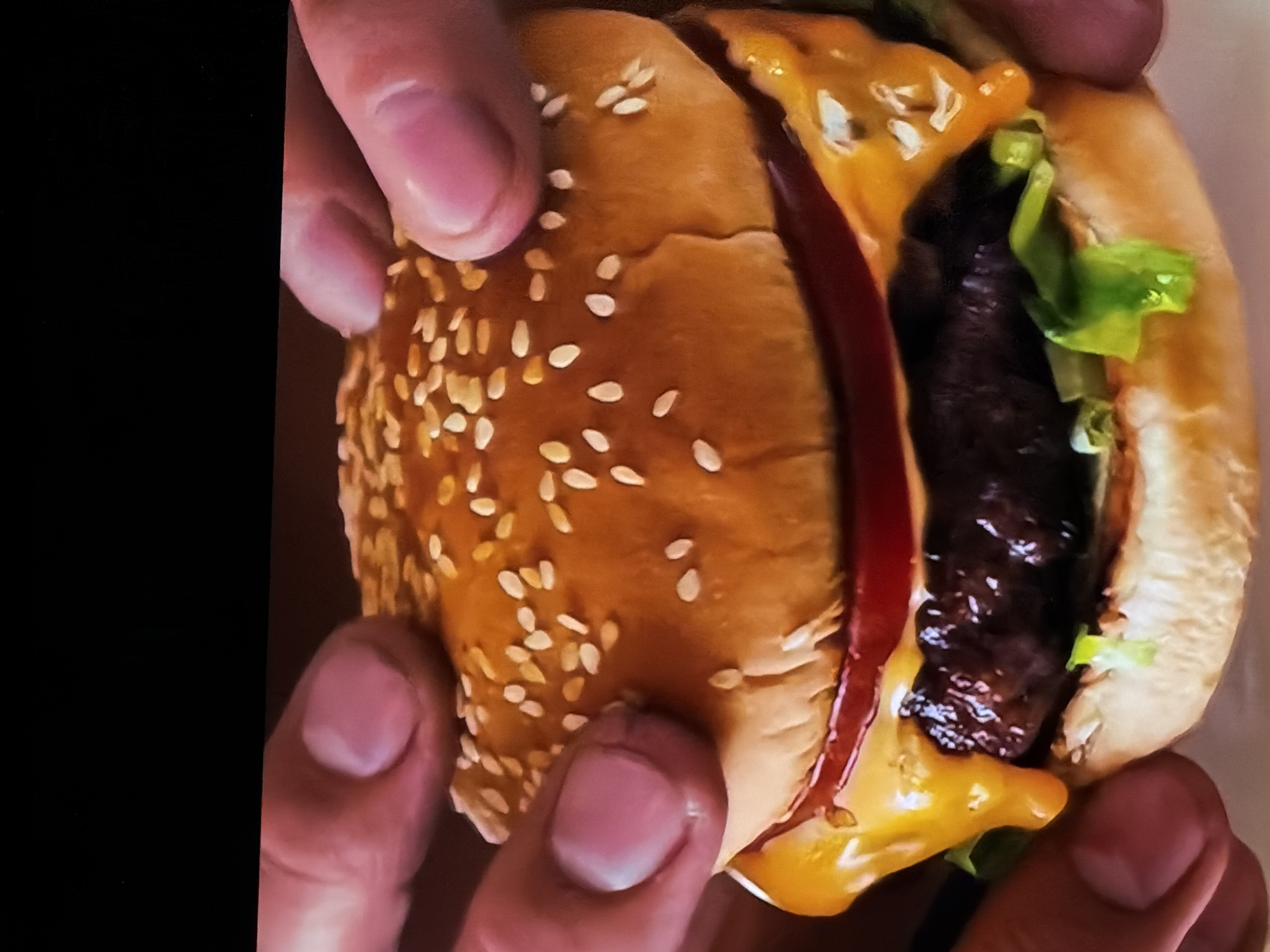 A close-up shot captures a person's hands holding a hamburger. The burger features a golden-brown sesame seed bun, which is gently gripped by fingers with a light skin tone and visible nails. Inside the bun, a thick, dark, and glossy meat patty is visible, generously topped with bright yellow, melted cheese. A strip of vibrant green lettuce peeks out from the right side of the burger, contrasting with a thin line of red sauce, possibly ketchup or a similar condiment, running along the left side of the patty. The hands appear to be presenting the burger or preparing to take a bite. The background is out of focus, with a dark, undifferentiated area on the left and a lighter, indistinct area on the right. The image conveys the appetizing nature of the food, set in the context of Sofiivska Borshchahivka, Ukraine. No text is visible.
