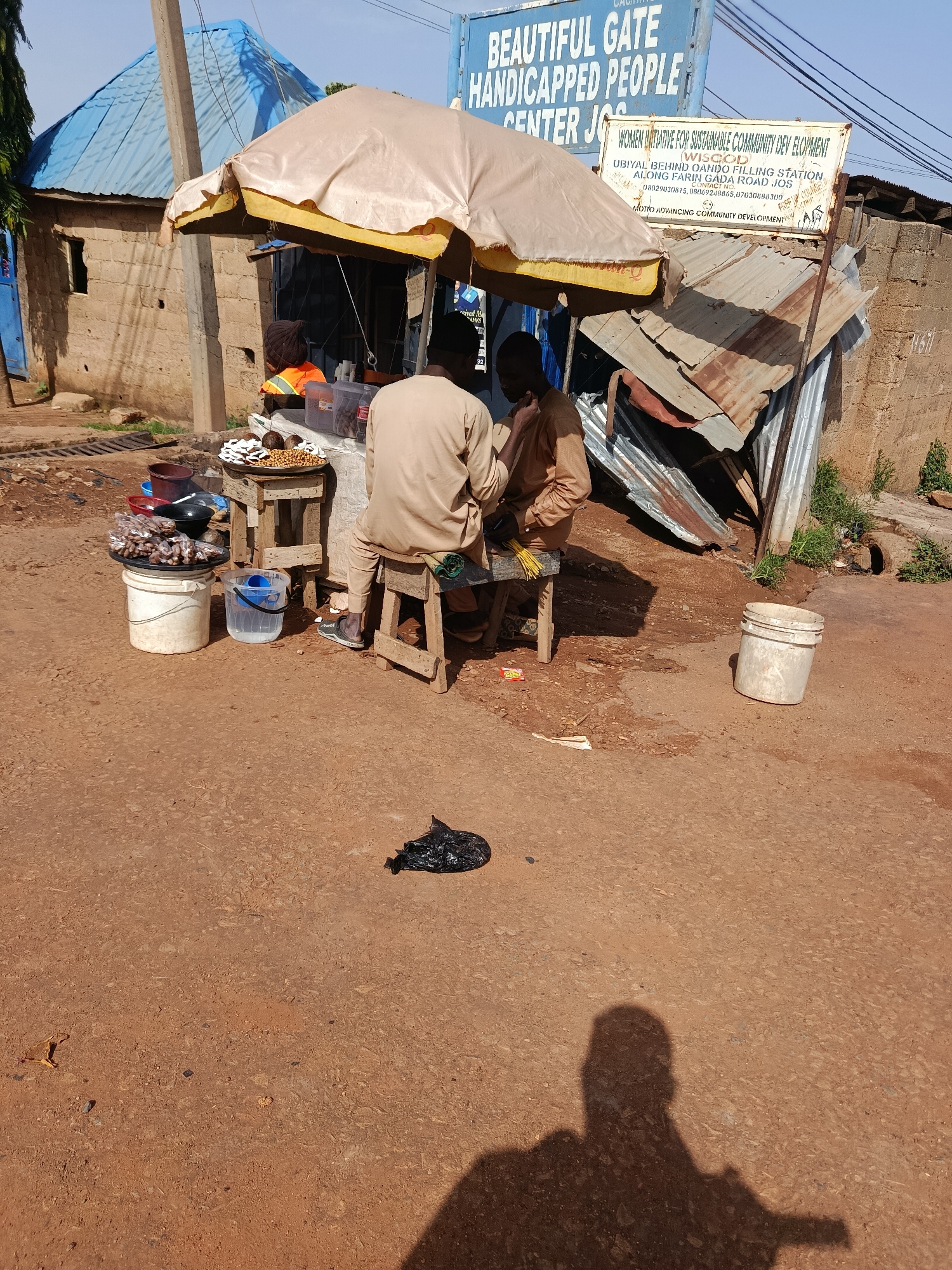 In Rafin Par, Nigeria, a street scene unfolds under a bright sky. Two men, dressed in traditional tan attire, sit at a makeshift stall. One man is seated on a wooden stool, facing away from the camera, while the other is on a bench, also facing away. The stall, covered by a large cream-colored umbrella, displays goods on wooden crates. On one crate, a pile of food items, possibly nuts and dried ingredients, is arranged on a plate. Another crate holds more food items, some appearing to be dried fish or meat, stacked in a black bowl. Two buckets, one white and one translucent blue, sit on the ground near the stall. A dark plastic bag is discarded on the red dirt road in the foreground. In the background, a building with a blue corrugated metal roof stands beside the stall. A blue sign with white lettering reads "BEAUTIFUL GATE HANDICAPPED PEOPLE CENTER JOS." Another sign, with white lettering on a white background, states "WOMEN INITIATIVE FOR SUSTAINABLE COMMUNITY DEV ELOPMENT (WISCOD)" and lists contact information and a motto. The overall impression is a bustling roadside market in a Nigerian city.