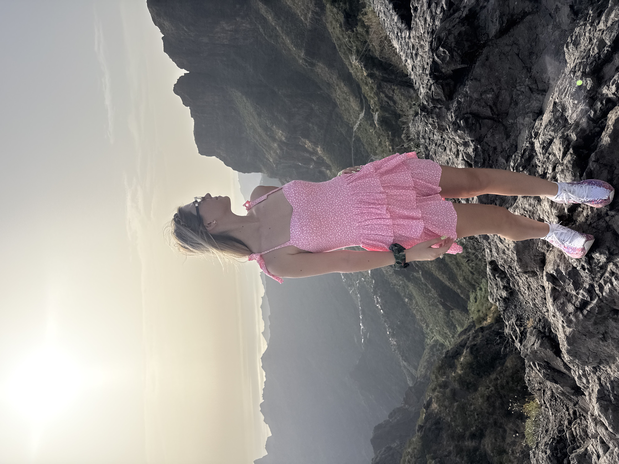 A young woman stands on a rocky outcrop overlooking a vast, mountainous valley. She is wearing a short pink dress with a floral pattern, a scrunchie on her wrist, and sneakers. The sun is setting or rising, casting a warm glow over the scene and creating a hazy atmosphere. The mountains in the background are rugged and steep, with a winding road visible in the distance. A small village is nestled in the valley below. The overall impression is one of natural beauty and solitude.
