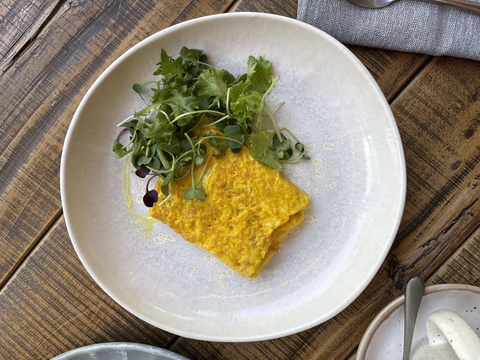 The image displays an overhead, static view of a meal set on a wooden table. A central, round, off-white ceramic plate with a speckled glaze holds a bright yellow, folded omelet with a visibly textured surface. Adjacent to the omelet, to its right, is a small heap of fresh green microgreens and frilly lettuce, interspersed with a few darker purple microgreens. A minimal amount of yellow liquid, possibly oil or dressing, is visible on the plate's surface near the omelet. The table surface is composed of dark brown, distressed wooden planks featuring prominent grain patterns and visible seams.
To the upper left of the main plate, a partial rim of another light-colored ceramic dish is visible. In the lower left quadrant, a portion of a speckled ceramic cup with a handle, a matching saucer, and a silver-colored spoon are partially present. To the lower right, a folded gray linen napkin and the tip of a metallic utensil are visible. The scene is illuminated by what appears to be natural light, casting soft shadows. There are no people present in the image. The specific location of Krasnoyarsk, Russia, cannot be determined from the visual information provided.