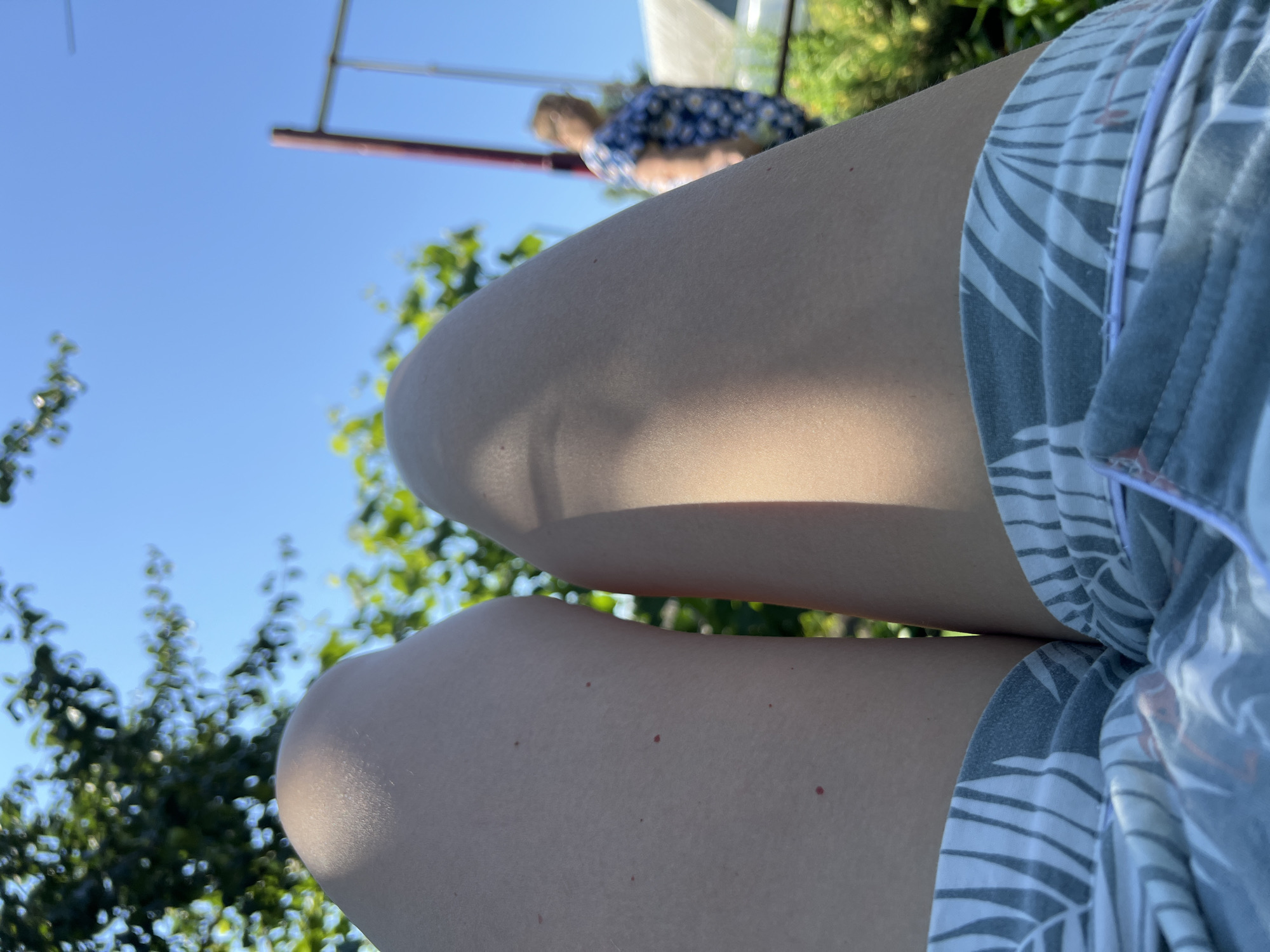 The image is a close-up, low-angle shot focusing on the legs of an individual wearing patterned shorts. The legs are positioned in the foreground, with the right leg slightly forward and casting a shadow from sunlight. In the background, out of focus, is a blue sky, lush green foliage, and a swing set with a person standing near it. The person in the background is wearing a blue and white floral-patterned dress. The environment suggests an outdoor, possibly a backyard or park setting, in Khryashchevka, Russia.