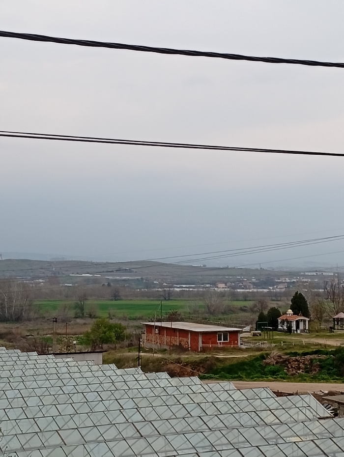 The image displays an outdoor scene in the city of Lebnitsa, Bulgaria, under an overcast, light grey sky. Two thick, dark electrical wires span horizontally across the top of the frame, with a thinner wire positioned below them. The lower third of the image is dominated by the roof of a large greenhouse complex, composed of numerous translucent or glass panels arranged in a grid pattern.
Beyond the greenhouse, in the midground, a rectangular, single-story building with reddish-brown brick walls and a flat, light-colored roof is visible, featuring at least one white-framed window. To its right, a small, white, chapel-like structure with a domed roof and a dark conical element stands, flanked by sparse vegetation and a wooden gazebo. The immediate ground around these structures consists of patches of green grass and bare earth. Additional power lines and poles are present in the mid-distance.
The background reveals a expansive rural landscape featuring green fields, fallow land, and scattered clusters of deciduous trees, many without foliage. Rolling hills are discernible in the far distance, merging with the uniform grey sky. No individuals or human activity are present in the scene.