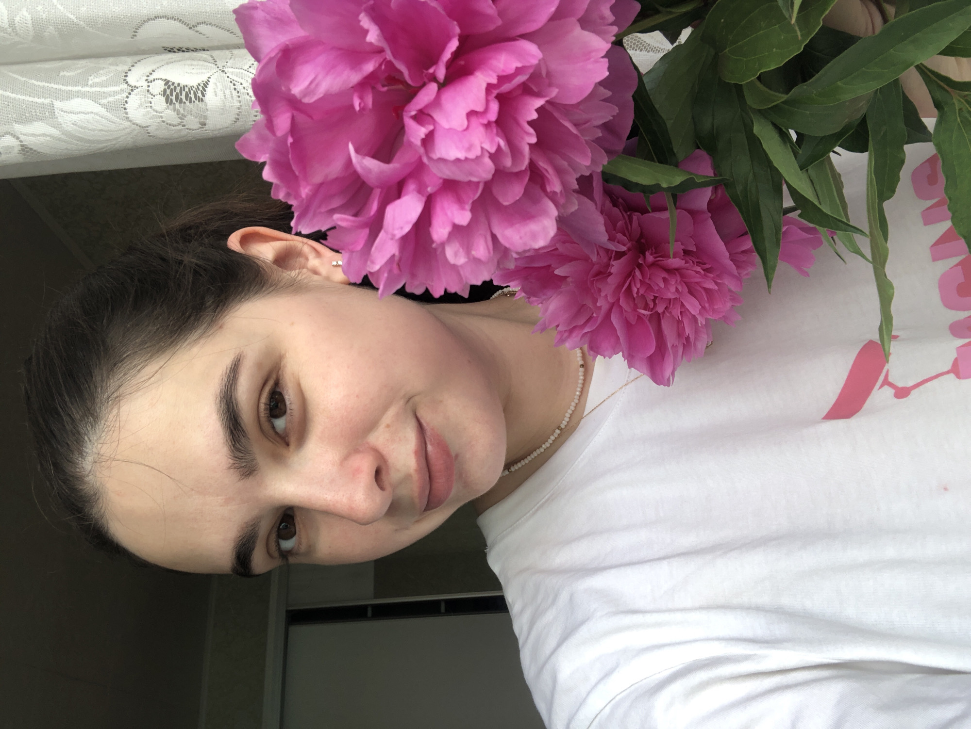 A young woman with dark hair pulled back and brown eyes poses for a selfie, holding a bouquet of vibrant pink peonies. She is wearing a white t-shirt adorned with a pink graphic design and a delicate beaded necklace. The background features a white lace curtain and a dark wall, suggesting an indoor setting, possibly a home in Noviy, Russia. The lighting appears natural, casting soft shadows. The overall mood is relaxed and cheerful.