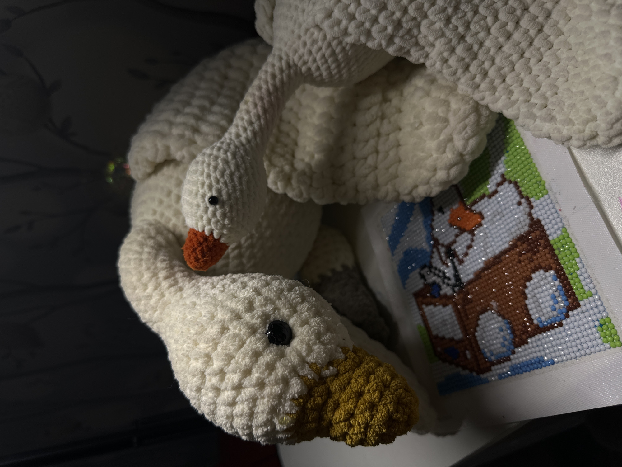The image depicts two stuffed, crocheted geese. One goose has an orange beak and the other has a yellow beak. The geese are white and appear to be lying on top of each other. In the background, there's a craft project made of small colored beads, featuring a depiction of a snowman. The setting is indoors with low lighting. It is impossible to determine if the location is in Chaikovsky, Russia, or anywhere else, based solely on this image.