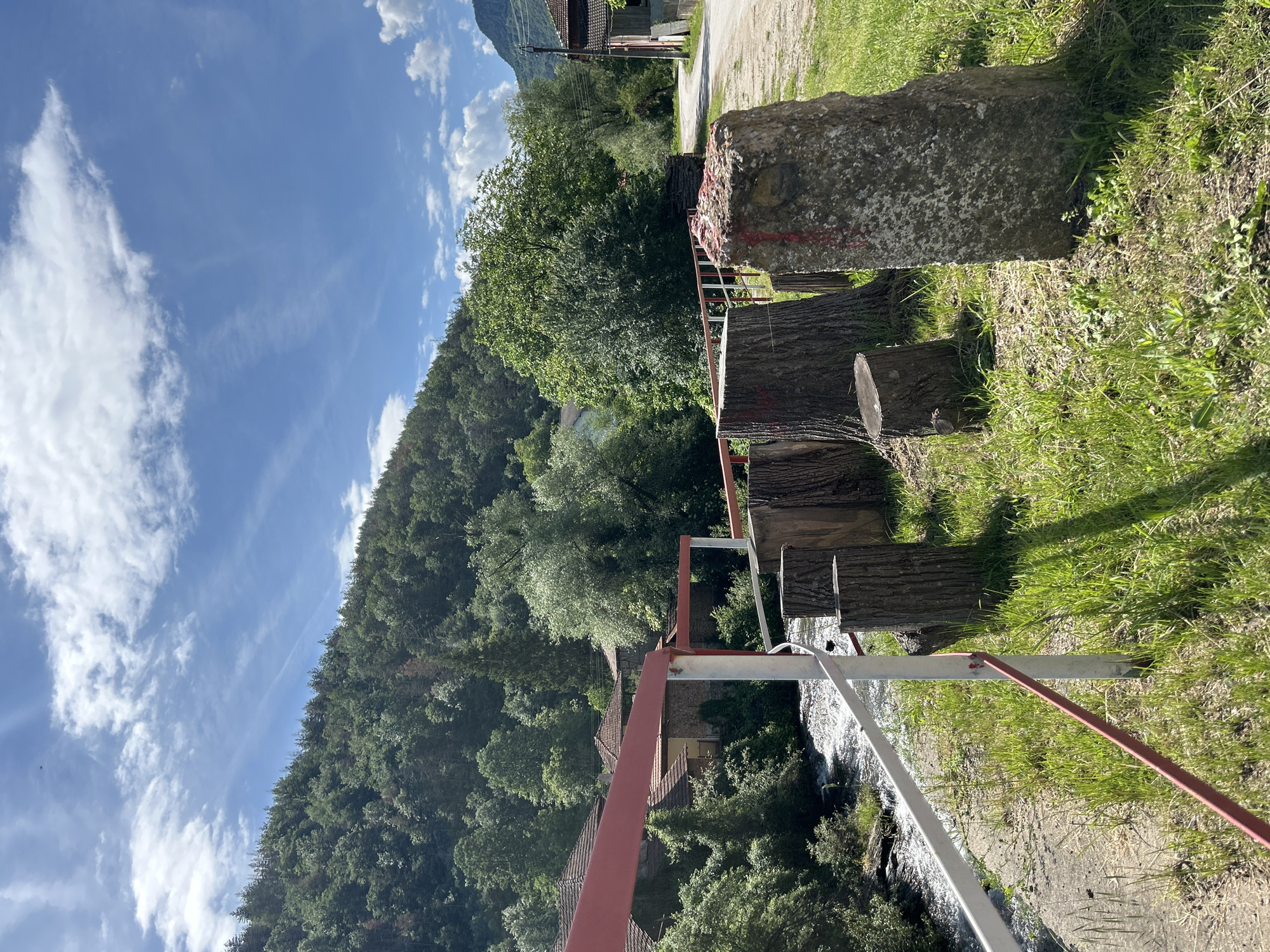The image captures a bright, sunny rural landscape with a mix of natural and man-made elements. The scene is dominated by a clear blue sky with scattered white cumulus and cirrus clouds. A densely forested mountain or steep hillside rises on the left and extends into the background.
In the foreground, on the right side, there are several large tree stumps and a prominent, rough concrete block. Both the concrete block and the tops of some stumps bear indistinct red markings, resembling lines or partial crosses. A red and white metal railing or bridge structure stretches horizontally across the mid-ground, with a fast-flowing stream or river visible beneath it on the left.
Further back, nestled among the trees on the right side of the frame, traditional-looking buildings with red-tiled roofs are discernible, suggesting a small village or Hamlet. A dirt road or path runs alongside the foreground elements on the right, curving into the distance.
The time of day is bright daylight, indicated by the strong sunlight and clear visibility, with noticeable shadows on the ground and objects. The weather is fair and sunny. No people are present in the image. The setting appears to be a verdant valley or mountainous region, showcasing a tranquil environment where human settlement coexists with natural forest and waterways.