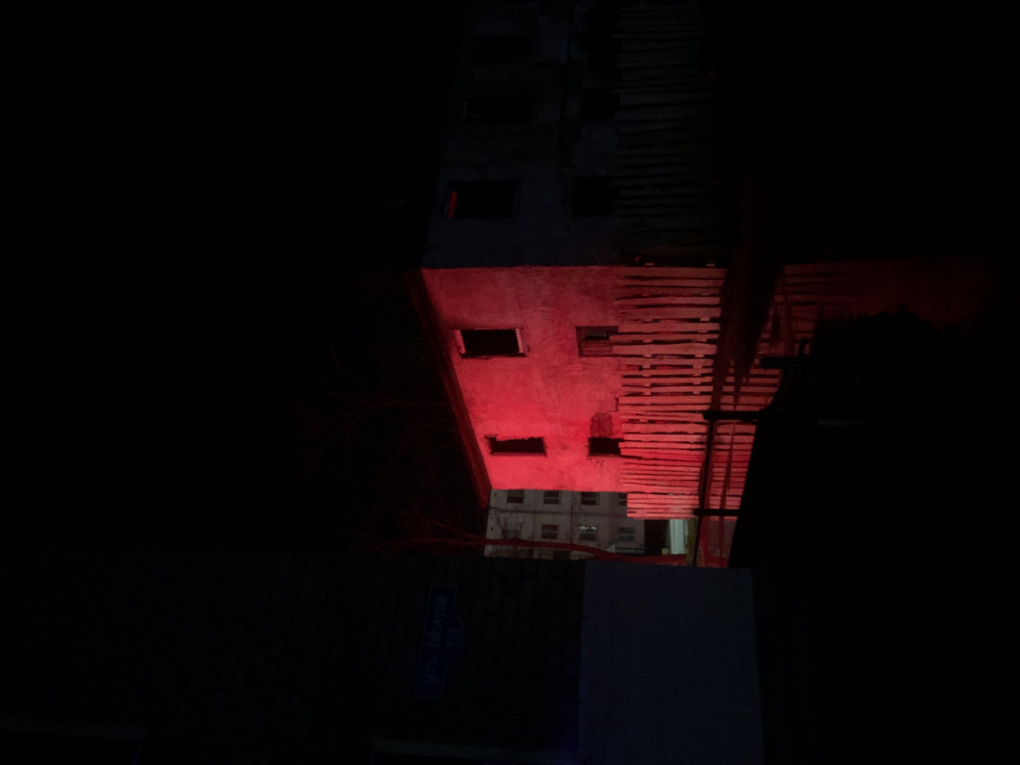 A low-angle, night-time photograph shows a street scene in what is likely Chaikovsky, Russia, with a partial view of a building illuminated in red. A street sign is visible on the lower left of the frame, reading "ул. Мира 19/10." The sign is blue with white text, and appears slightly blurred. Above the street sign is a portion of a building's façade, marked by several square window openings. This section is illuminated in a strong red light. To the right of the façade, horizontal wooden slats are visible, also illuminated in red. In the distance, above the first building, a lighter, indistinct building with multiple windows can be seen. The overall impression is of a dark urban environment, with the intense red light drawing the viewer's eye. The rest of the scene is dark, obscuring details.