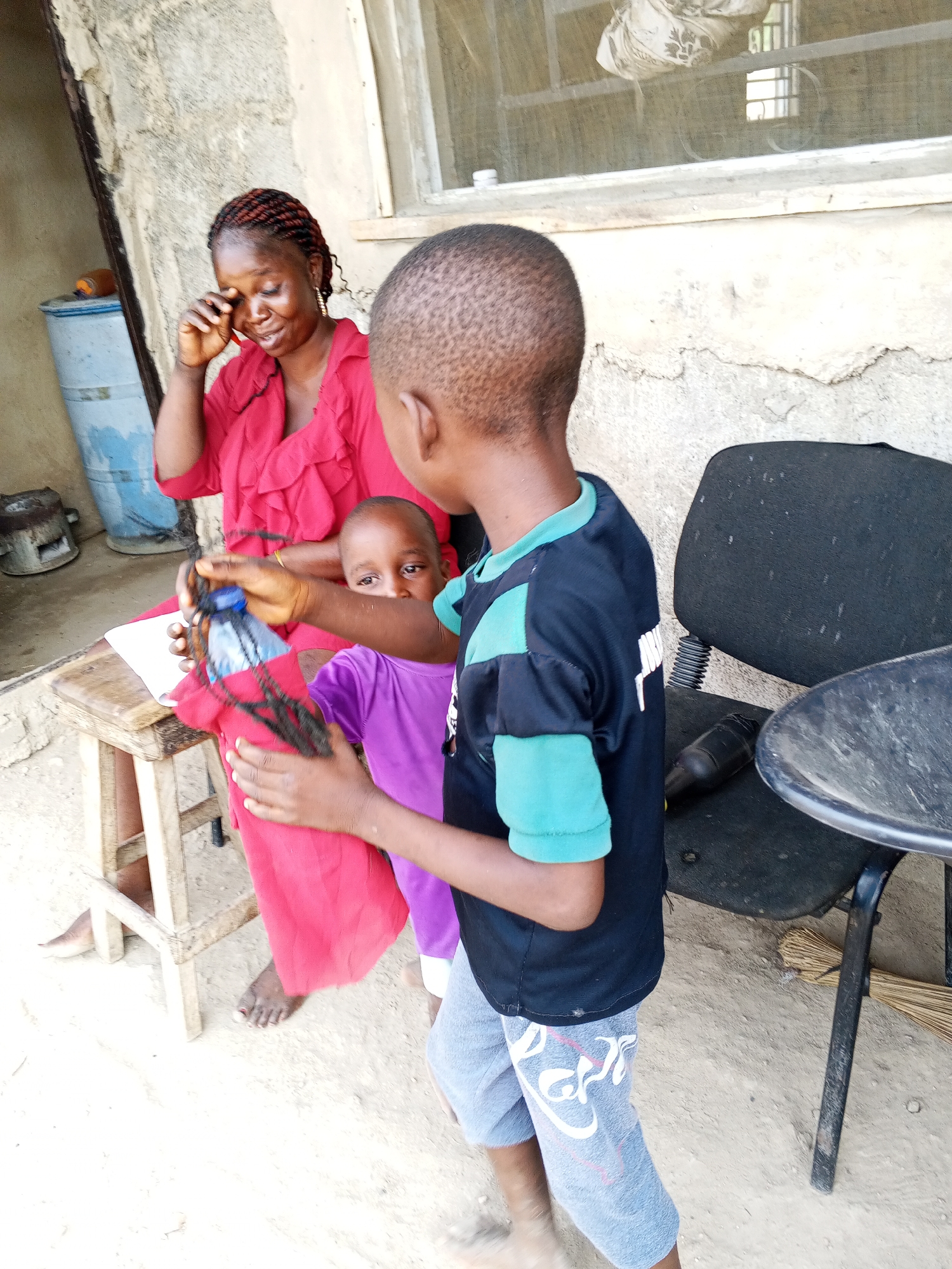 A medium indoor/outdoor shot in the city of Unknown, Unknown shows a woman in a red top sitting on a wooden stool; she is holding a water bottle covered with dark hair in her right hand. Behind the woman is a blue barrel, and next to it, what appears to be a cooking implement. The woman is holding her left hand to her face as if she is wiping away tears, but she is smiling. Next to her, a smaller child in a purple shirt is looking at the camera with curiosity. A boy in a black and blue shirt, with grey pants, stands in front of them, facing away from the camera, perhaps interacting with the others. To the right of the children is a black office chair and a small, round table. Behind the woman is a wall with a window. The floor appears to be dirt or sand.