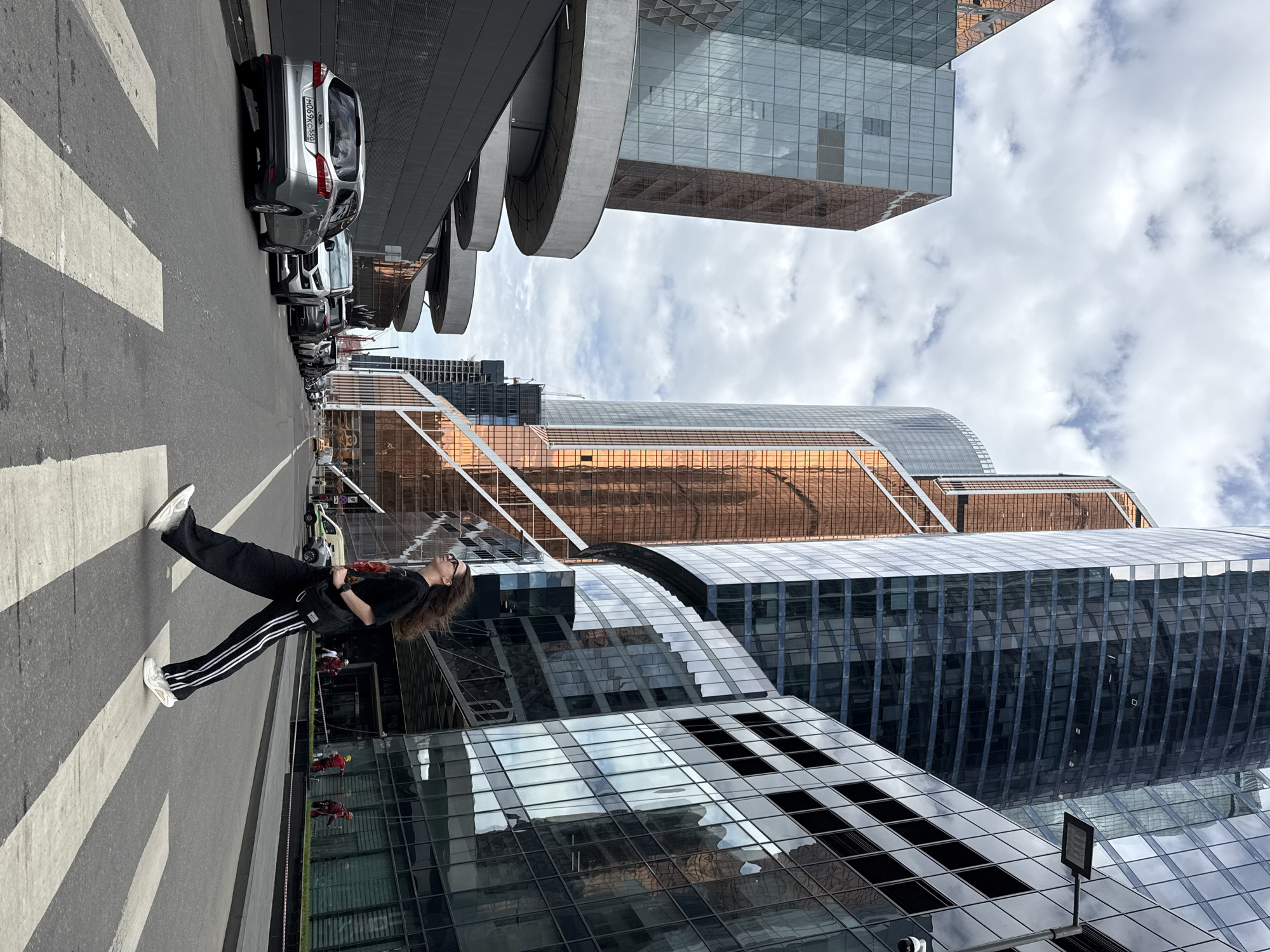 A woman in black clothing with white stripes on the sides of her pants lies on a pedestrian crosswalk in a city environment. The background features modern architecture, including skyscrapers with reflective glass exteriors. Several cars are parked alongside the street. The scene appears to be in the city of Khimki, Russia.