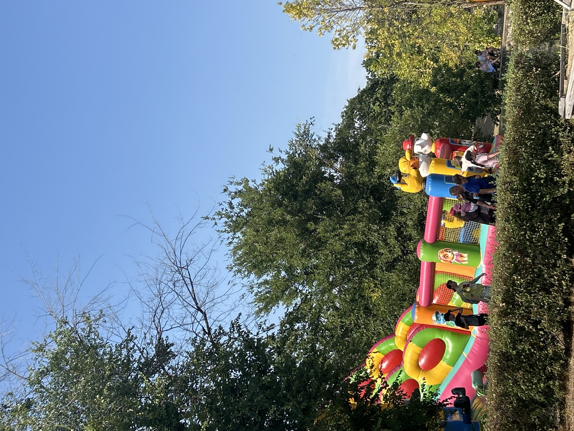 A bright, inflatable playground with multiple slides and cartoon characters is set up outdoors amidst lush green trees. Several children and adults are present, some appearing to be entering or exiting the inflatable structure, while others are standing nearby. In the background, a few people are seated on a stone ledge. The scene is under a clear blue sky on a sunny day. This location is in the city of Pivdennoukrainsk, Ukraine.