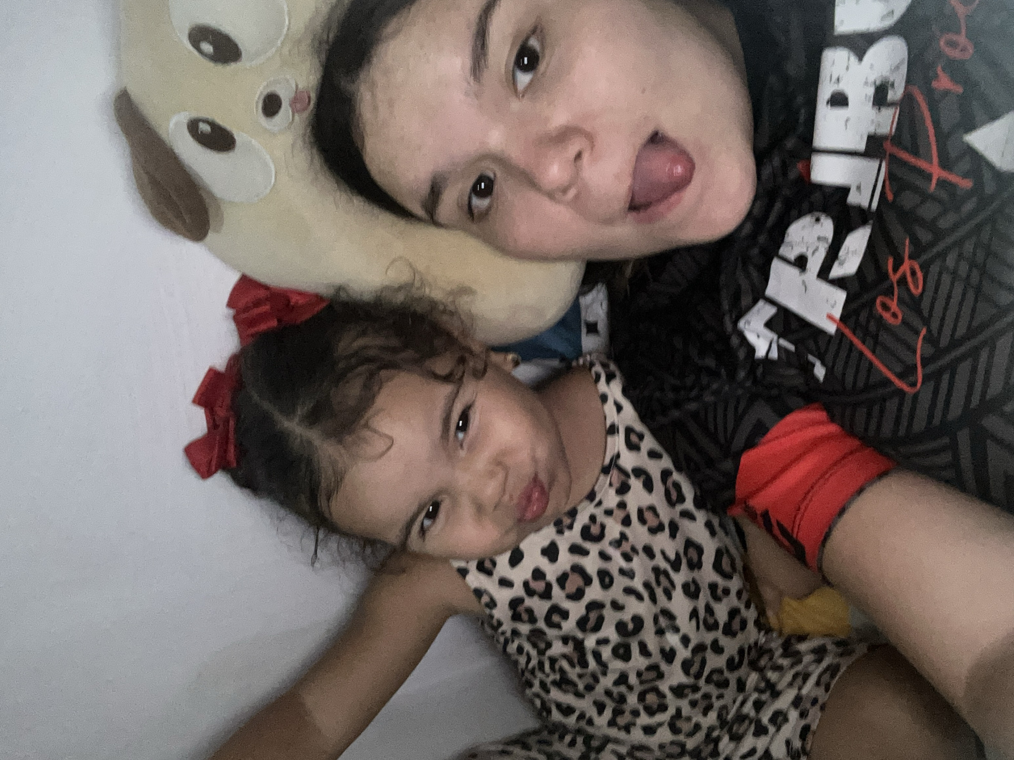 The image shows a selfie taken indoors, likely in a residential setting. Two individuals are visible: an adult female and a young female child. The adult is making a face with her tongue sticking out, while the child is pouting. The child is wearing a leopard-print dress with a red bow in her hair. The adult is wearing a dark shirt with a graphic print. A stuffed animal is positioned near the adult's head. The background is a plain white wall. No elements are visible that indicate the location is San Juan de Los Morros, Venezuela.