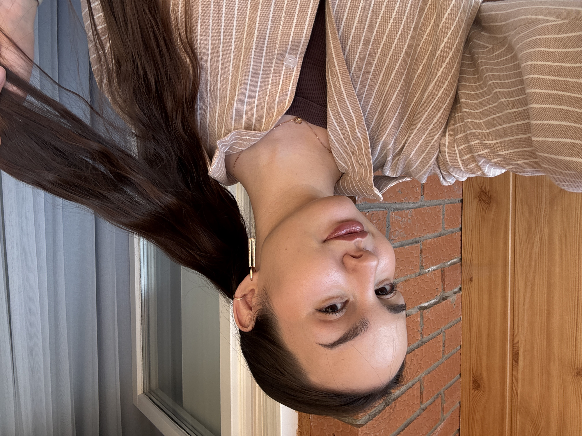 A woman poses in front of a window covered by sheer curtains and a brick wall adjacent to a wooden door. She is wearing a tan striped shirt over a dark brown top. Her hair is long and brown, and she wears gold rectangular earrings. The photo was taken indoors. It is not possible to identify the location as Chaikovsky, Russia, based on the image.