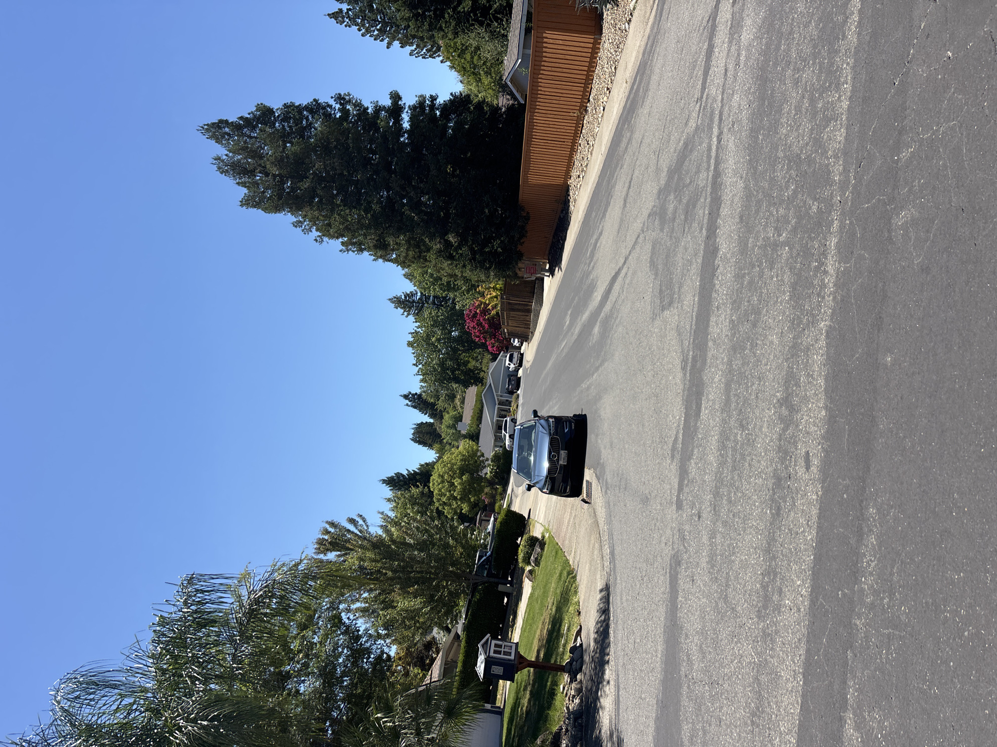 The image captures a suburban street scene on a clear, sunny day in Folsom, California, United States of America. A dark-colored Volvo SUV is parked on the right side of the asphalt road, angled slightly towards the viewer. The street curves gently to the right, lined with residential homes and mature trees on both sides. On the left side of the road, a small blue and white mailbox stands on a post in a grassy area. The houses visible have various architectural styles, with some featuring pitched roofs and wood or stucco exteriors. A tall, dark wooden fence runs along the right side of the street. Lush green lawns and trees, including palm trees and tall evergreens, contribute to the neighborhood's appearance. The sky is a vibrant, cloudless blue.
