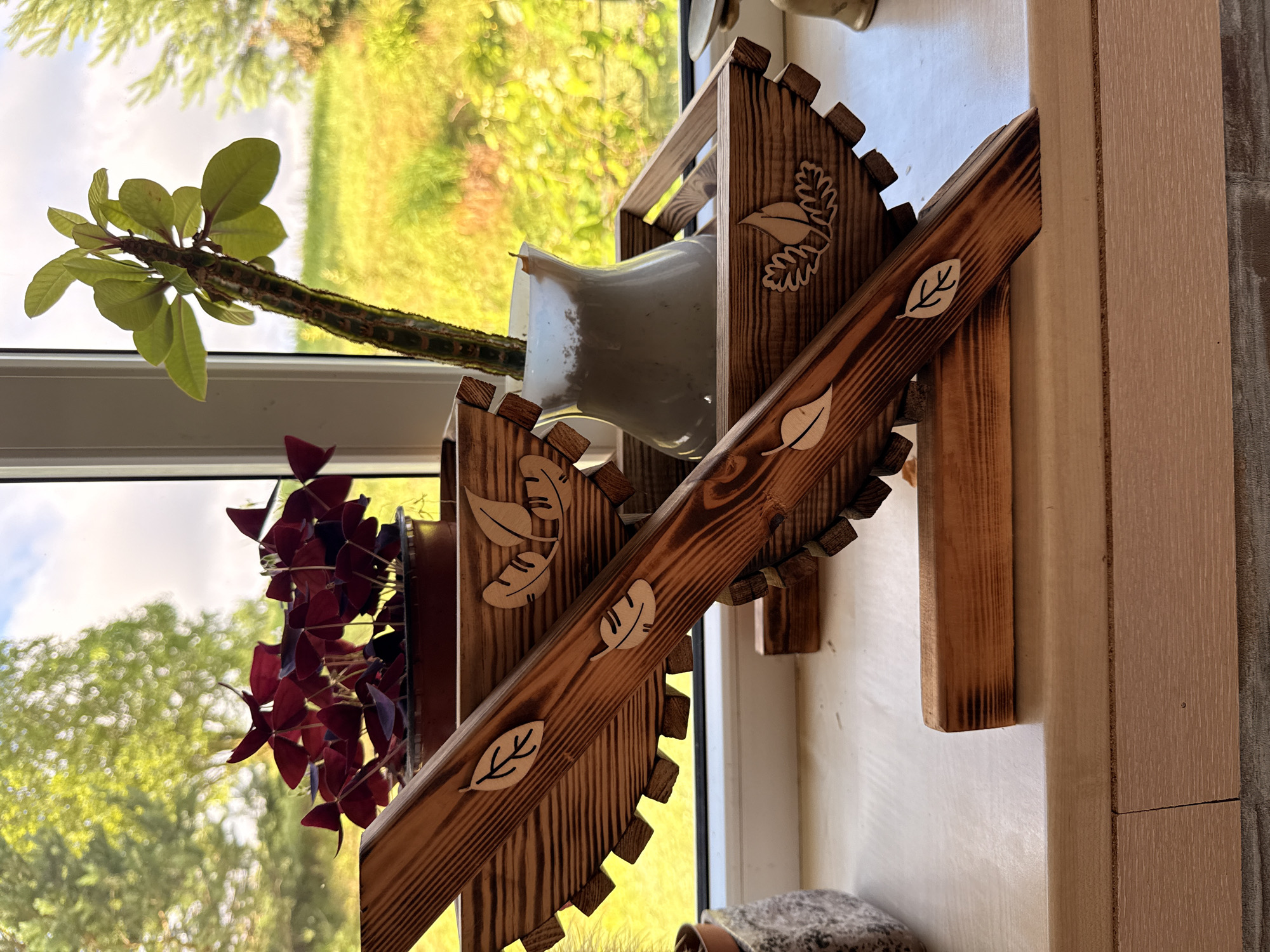 The image depicts a windowsill in a home in Galinė, Lithuania, with a view of greenery outside. On the windowsill, there are two wooden plant stands. The foreground stand is made of dark, stained wood with lighter wood leaf cutouts and sits at an angle. Behind it, a second wooden stand holds a white vase containing a plant with green leaves and a mottled stem. To the left of the stands is a potted plant with dark red, triangular leaves. The windowsill itself is light-colored wood, and the background shows a blurred natural landscape with trees and grass under a cloudy sky.