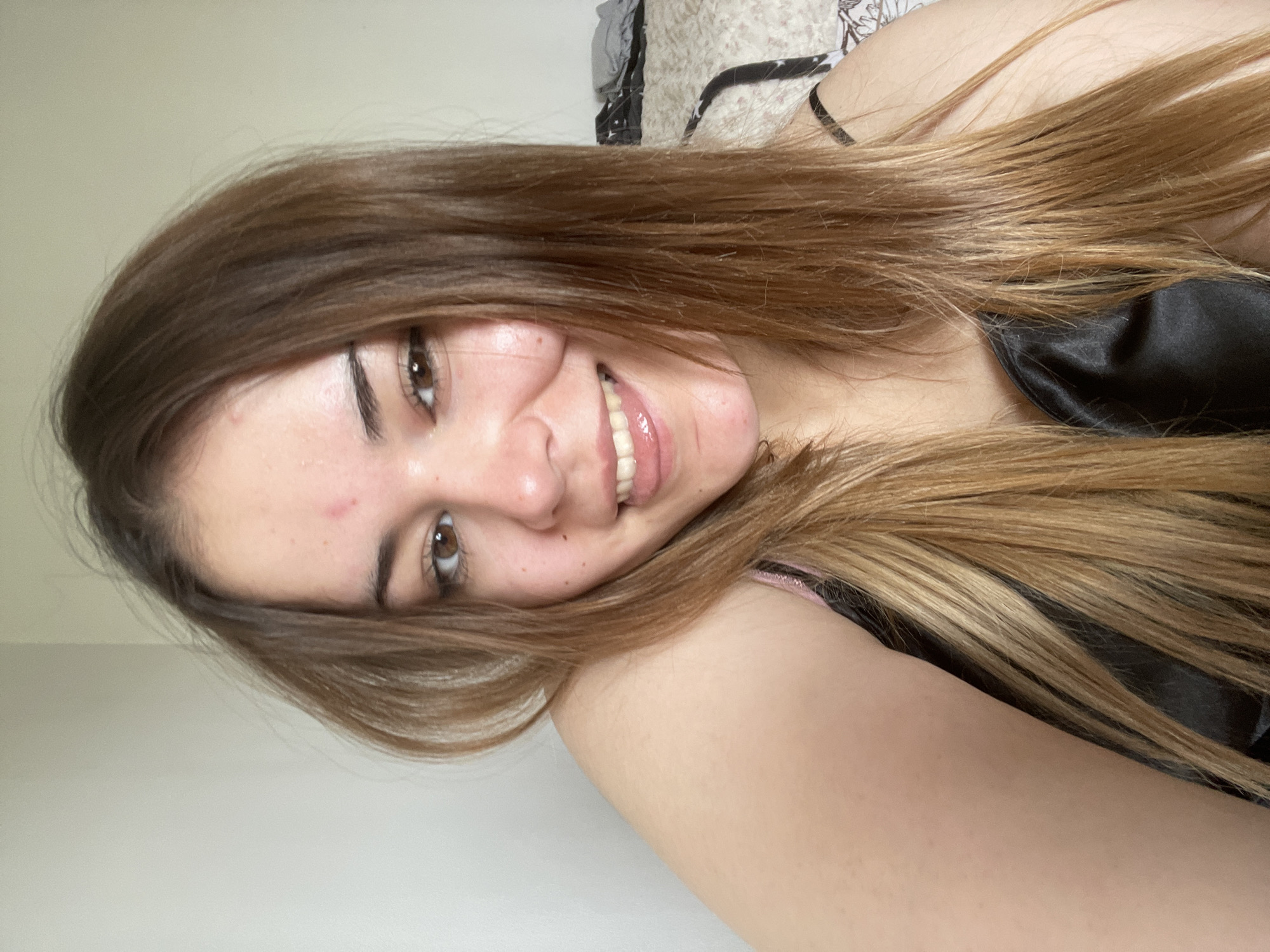 A young woman with shoulder-length brown and blonde hair is smiling at the camera. She is wearing a black silky top. The background is a cream-colored wall and part of a bed with patterned bedding. The location is not identifiable as Plovdiv, Bulgaria based on the provided image.