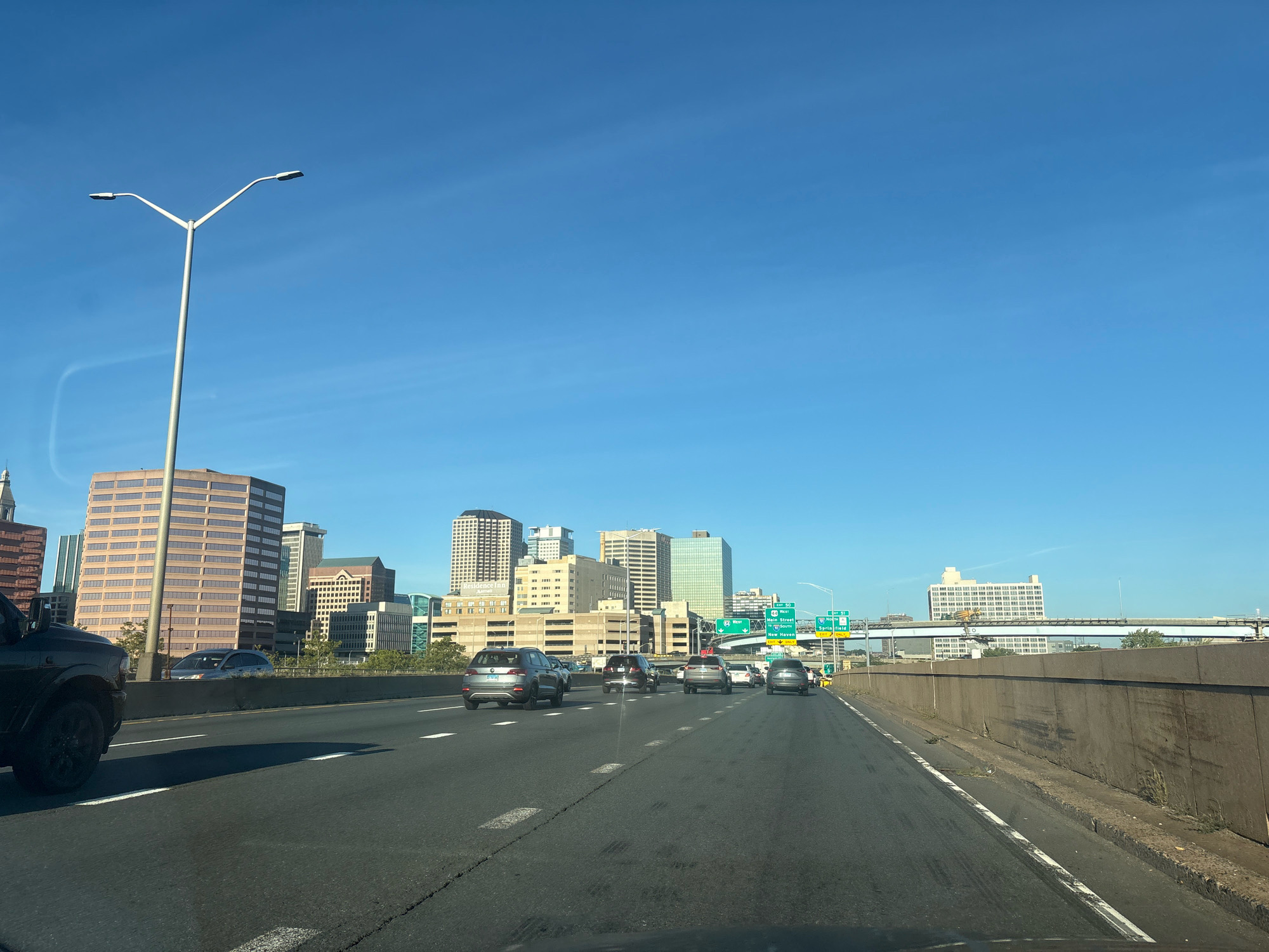 The image shows a view from a vehicle on a highway approaching the city of Hartford, Connecticut, United States of America. The foreground features the road surface with lane markings and a concrete barrier on the right. Several cars are driving on the highway. In the middle ground and background, a skyline of various office buildings is visible, characterized by a mix of modern and older architectural styles. A tall streetlamp with two lights is positioned on the left side of the image, against a clear blue sky. Highway signs indicating exits for "Main Street," "New Haven," and "Springfield" are present. The environment appears to be daytime with sunny weather.