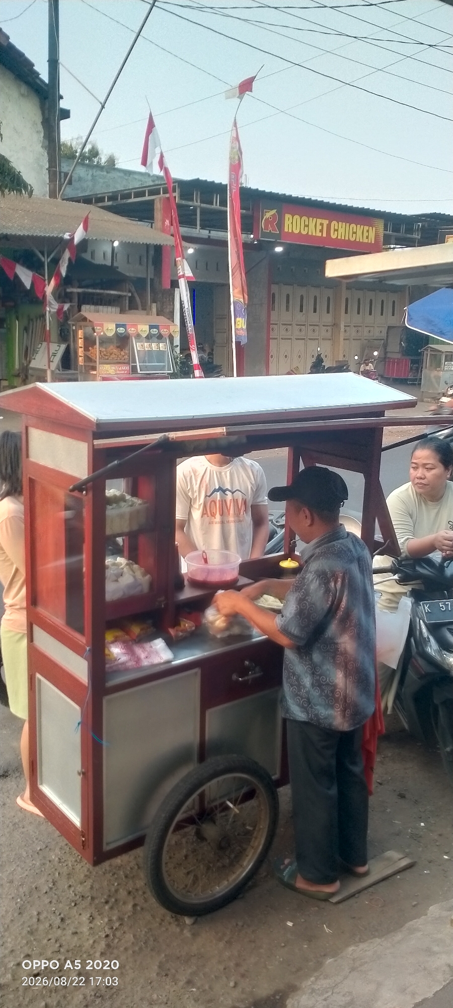 This is a full-shot, eye-level, outdoor, vertical photograph taken on a street in Kedung, Indonesia. In the foreground, a food cart is the main subject. The cart is made of dark wood with a white countertop and has glass doors displaying food items. A man wearing a patterned blue shirt and black pants is standing behind the cart, preparing food. He is wearing a black baseball cap and is holding a plastic bag with food. A woman in a light-colored t-shirt is seated to the right of the cart, looking towards the food preparation. Another woman in a light-colored top and a light-colored skirt is standing to the left of the cart, partially visible. In the background, there is a building with a red sign that reads "ROCKET CHICKEN." Several Indonesian flags are visible, attached to poles and hanging from wires. The ground is paved with rough dirt and some scattered debris. The sky is visible at the top of the image and is a light blue color. The overall scene suggests a street food vendor operating in a commercial area.