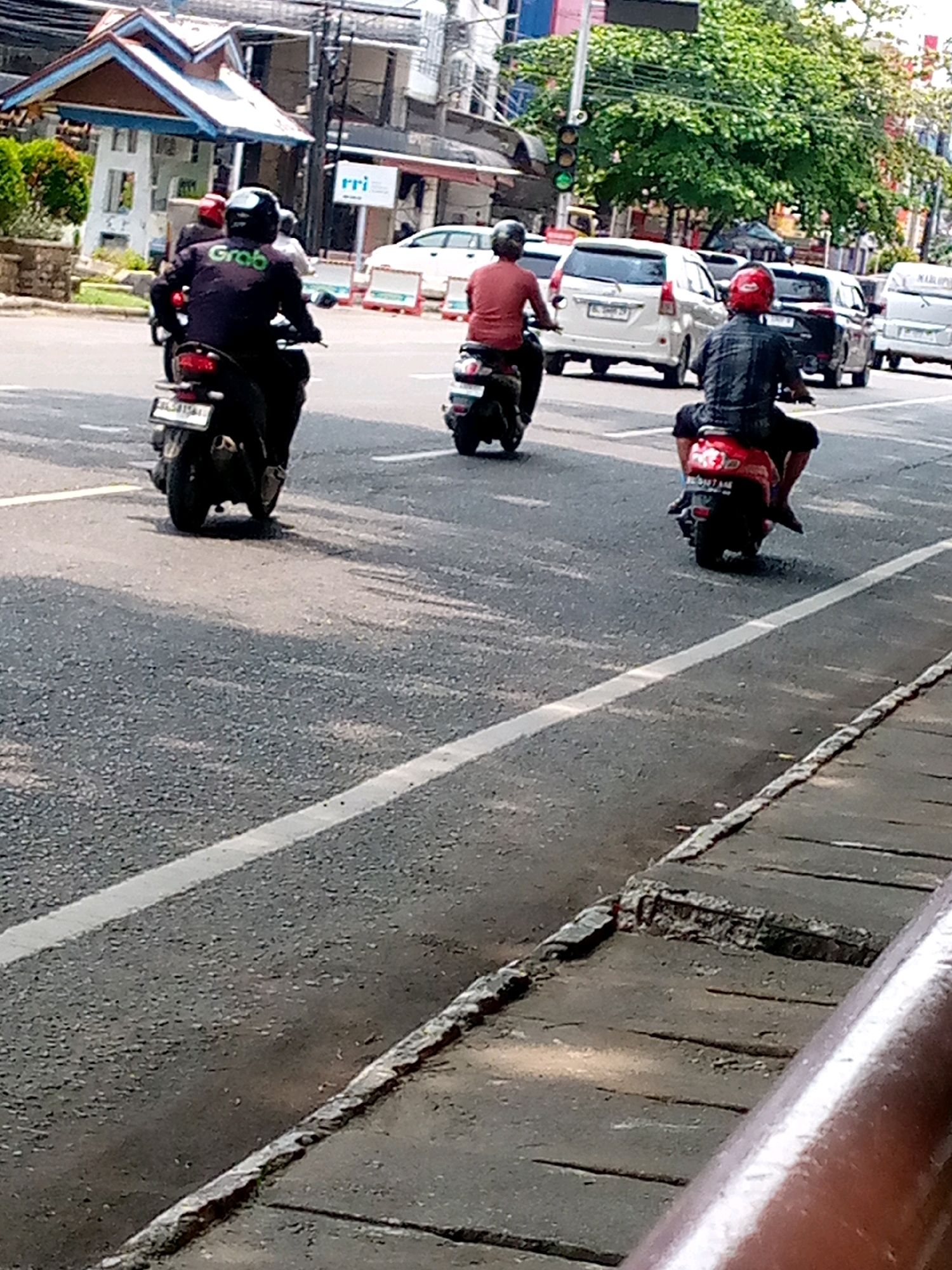 The image shows a street scene in Lhokseumawe, Indonesia. Three people are riding motorcycles. The person in the foreground on the left is wearing a dark jacket with "Grab" written on the back, a black helmet, and is riding a black motorcycle. The person in the middle is wearing a red shirt and a helmet, riding a scooter. The person in the foreground on the right is wearing a checkered shirt and a red helmet, riding a red scooter. Multiple cars are also visible on the road, including white and black vehicles. Buildings and trees line the street, and traffic lights are present. The road surface is asphalt with white lane markings. The scene appears to be a typical urban traffic environment.