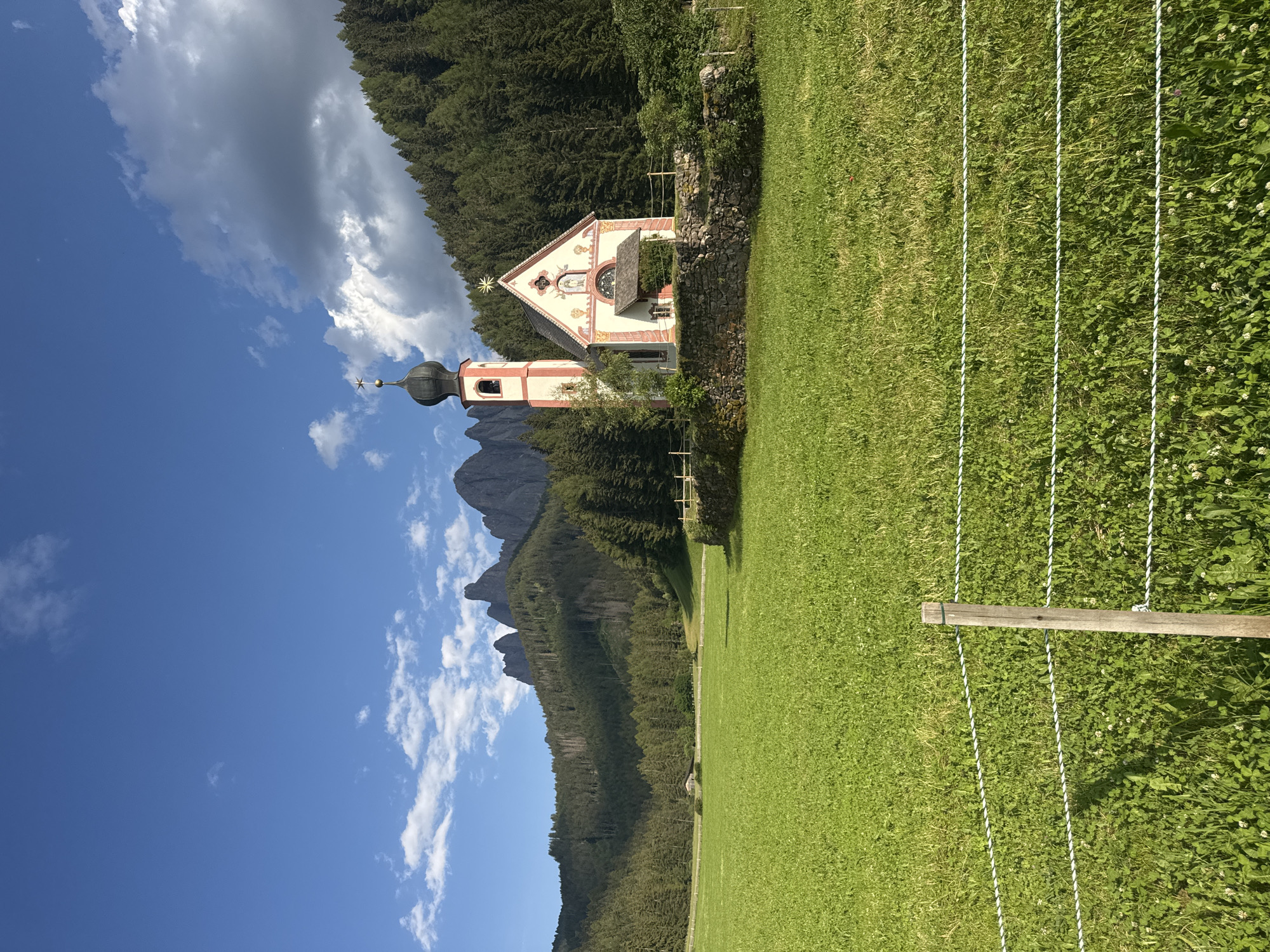 This is a vertically oriented, eye-level shot of the St. Magdalena church in the Italian Alps. The church, with its characteristic red and white striped bell tower and ornate facade, is nestled against a backdrop of dense evergreen trees and dramatic, jagged mountain peaks. A bright blue sky with scattered white clouds dominates the upper left portion of the image. In the foreground, a vibrantly green, well-maintained grassy meadow stretches across the right side, separated from the church and trees by a low, rustic stone wall. Several strands of white, twisted rope, supported by a wooden post, run horizontally across the meadow, suggesting a boundary or perhaps part of a temporary fence. The sunlight suggests it is daytime, likely mid-morning or afternoon, casting subtle shadows and illuminating the landscape. The overall impression is one of serene beauty and picturesque alpine scenery. No people or text are visible.