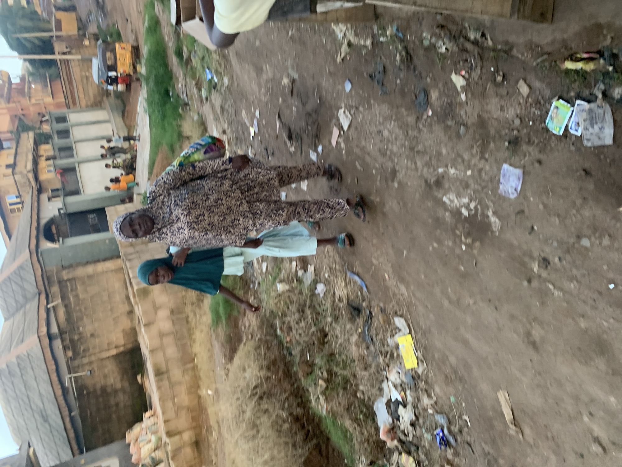 In Shagamu, Nigeria, a woman and a young girl walk down a dirt path lined with debris and vegetation. The woman, wearing a patterned dress and headscarf, carries a colorful bag. The girl, in a green headscarf and light blue dress, walks beside her. In the background, buildings and a yellow auto-rickshaw are visible, suggesting a semi-urban environment. The ground is uneven and littered with various objects, indicating a lack of formal waste management. The scene captures a moment of daily life in a developing area.