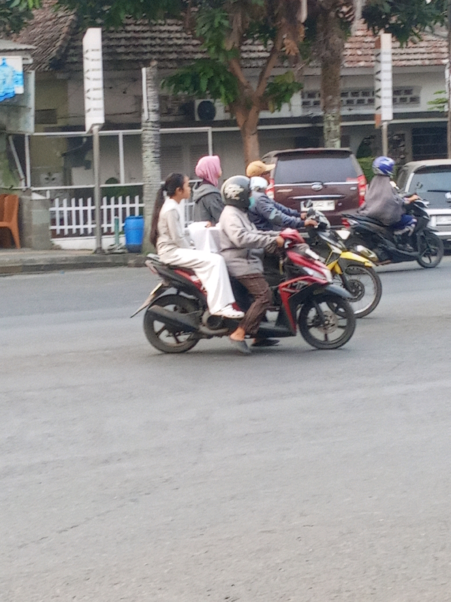 In Lubuklinggau, Indonesia, a group of people are riding motorcycles on a street. In the foreground, a person wearing white clothing and a pink headscarf rides a black motorcycle, carrying a passenger also in white. Behind them, another person in a grey jacket and helmet is on a red motorcycle. Further in the background, a person in a grey jacket and blue helmet is on a black motorcycle, followed by another motorcycle. A maroon car is visible behind the motorcycles. The street is paved, and there are buildings and trees on the side of the road.
