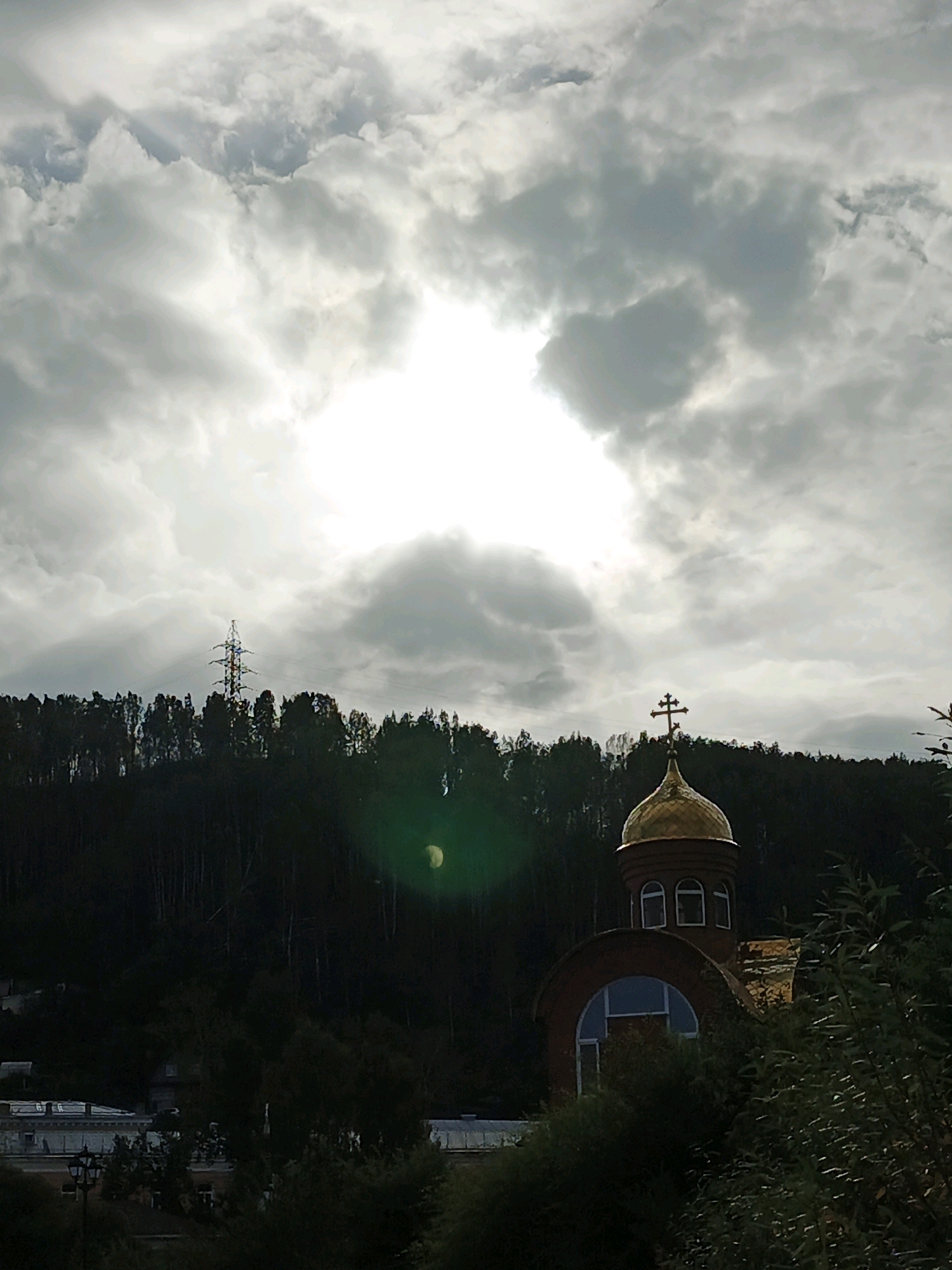 The image depicts a panoramic view of a residential area on a hillside in Zlatoust, Russia, under a dramatic, overcast sky. In the foreground, lush green foliage partially obscures a brick building with a golden dome, which appears to be part of an Orthodox church. The building features arched windows and a cross atop the dome. Behind the church, a dense forest covers the hillside, with a tall, lattice-like electrical tower visible in the distance, silhouetted against the bright, diffused sunlight breaking through the clouds. The lower part of the image shows a few low-rise buildings and streetlights, suggesting a street or park area. The overall atmosphere is somber yet serene, with the natural landscape dominating the scene.