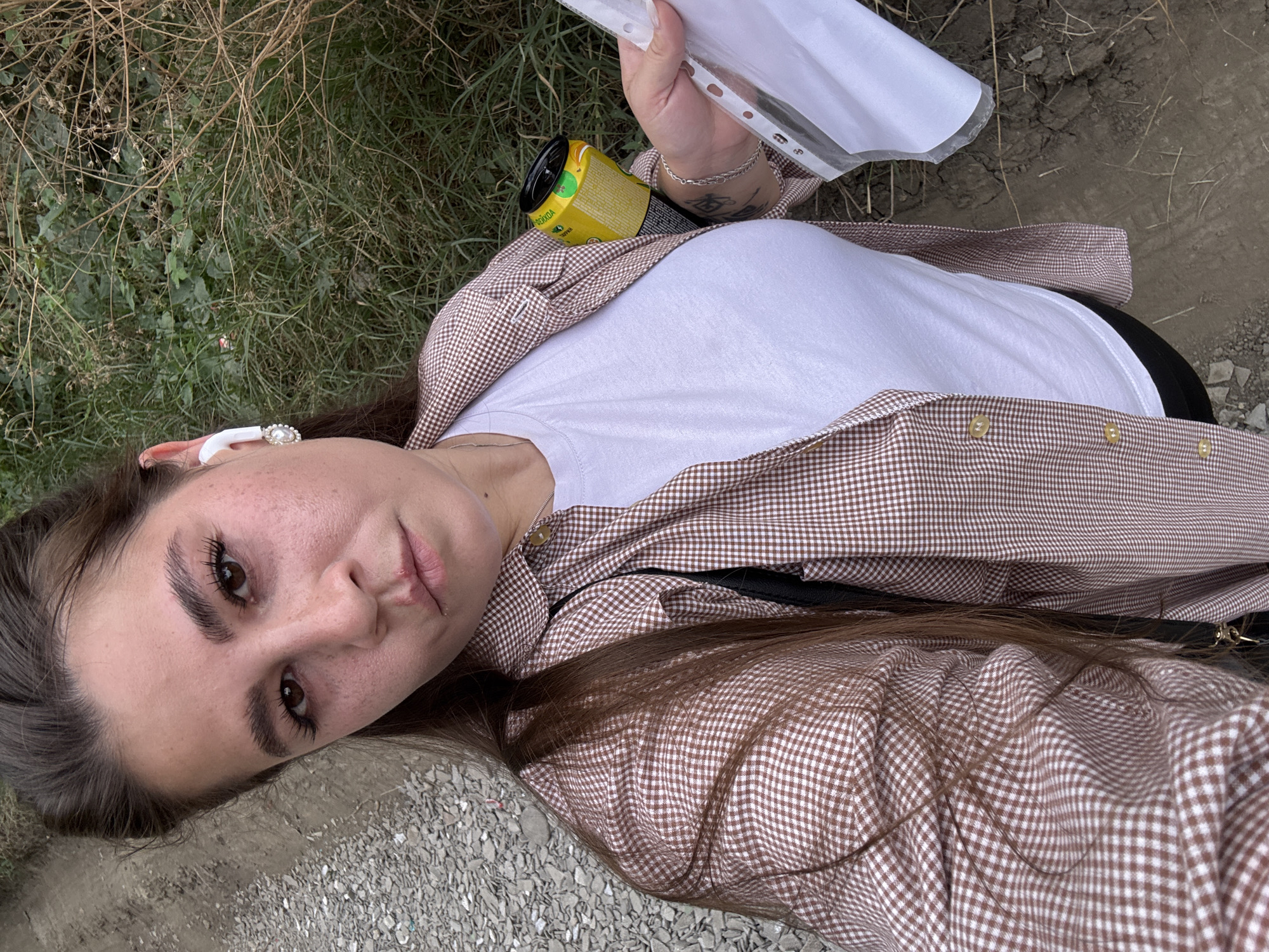 A woman with long brown hair is taking a selfie outdoors. She is wearing a white t-shirt and a brown and white checkered shirt. She has a white earbud in her left ear and is holding a yellow can and a clear plastic sleeve with papers. The background appears to be a rural or undeveloped area with gravel, dirt, and dry vegetation. The woman has a split lip and some redness on her cheeks. The location is identified as Nadezhda, Russia.