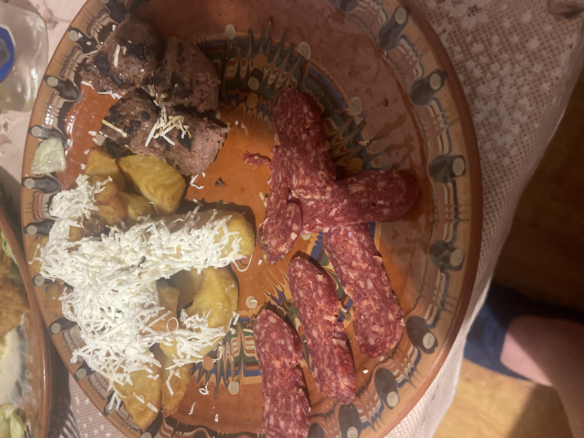An overhead shot captures a rustic meal served on a round, terracotta-colored earthenware plate adorned with traditional, colorful geometric patterns around its rim. The plate holds an assortment of food items.
In the upper left, there are several pieces of grilled meat, possibly small meatballs or kebabs, with some charred marks. A sparse scattering of shredded white cheese or similar topping is visible on these. To the left of the meat, a small pile of sliced, light-colored vegetables, possibly cucumber, is present.
Dominating the lower-left and central portion of the plate are golden-brown fried potato wedges, heavily blanketed with a generous amount of shredded white cheese. On the right side of the plate, several elongated pieces of sliced cured sausage are arranged. These sausages appear reddish-pink with visible specks of fat and spices.
The setting appears to be indoors, likely during a meal. A white tablecloth with a subtle woven pattern is visible beneath and around the plate. A partial view of a clear bottle, possibly containing water or a beverage, is seen in the upper left corner. In the bottom right, a person's leg and foot, clad in dark blue fabric, are partially visible, suggesting someone is seated at the table. The lighting suggests an indoor, possibly evening, setting with artificial light. No discernible text is visible.